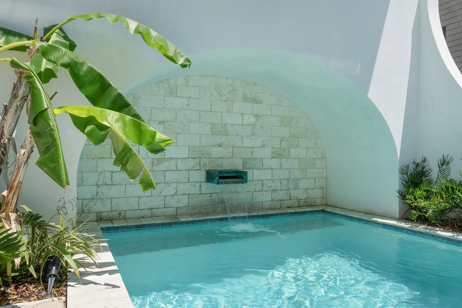 Private backyard swimming pool with a waterfall feature on a white brick wall, surrounded by tropical plants, including banana leaves and palms.