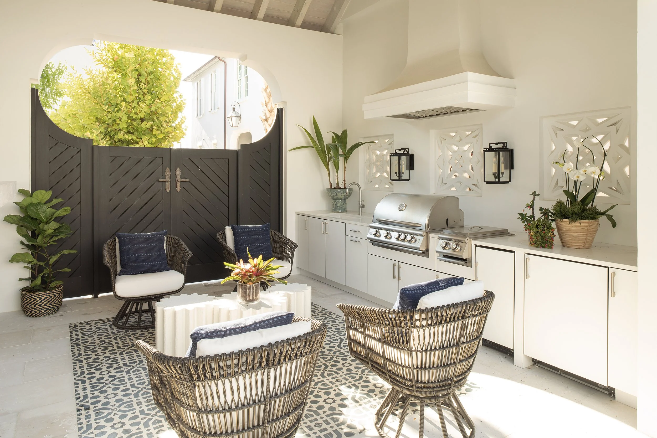 Outdoor kitchen and seating area with wicker chairs, a white countertop, potted plants, and black and white patterned tiles, with a black fence and green trees in the background.