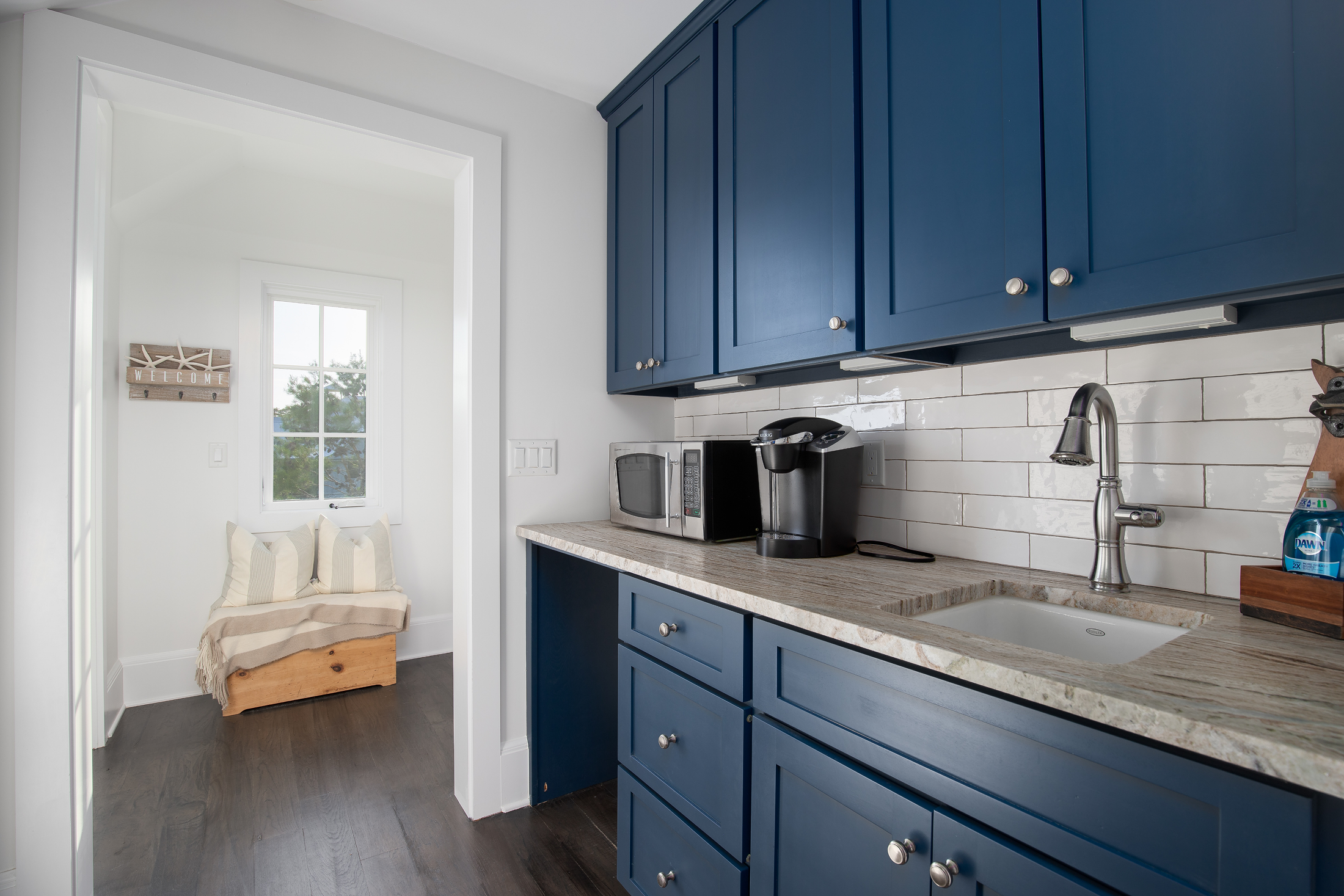 Modern kitchen with blue cabinets, a beige stone countertop, a stainless steel sink with a faucet, and small kitchen appliances including a microwave and coffee maker. There is a doorway leading to a bright sitting area with a window and a wooden ben