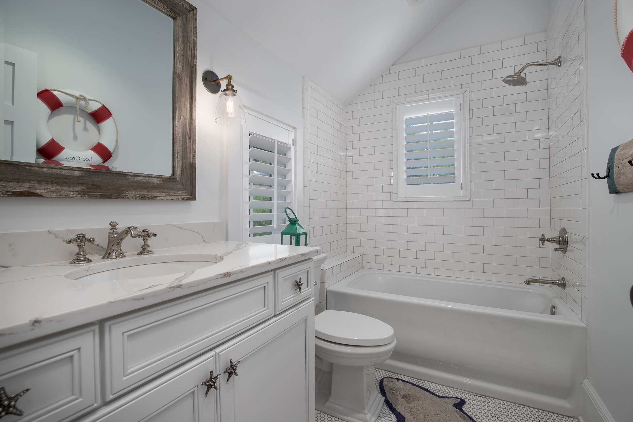 Bathroom with white vanity, marble countertop, wall mirror, toilet, white tiled bathtub area, window with shutters, and nautical decor.