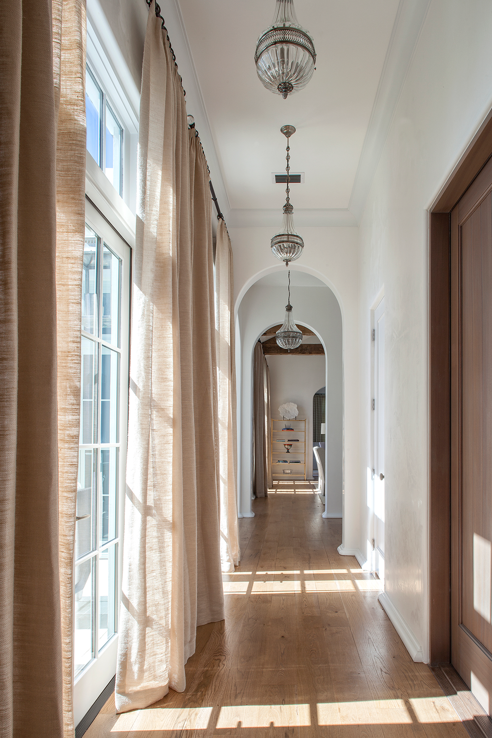 Bright hallway with wooden floors, beige curtains, and three chandeliers hanging from the ceiling, leading to a room at the end.