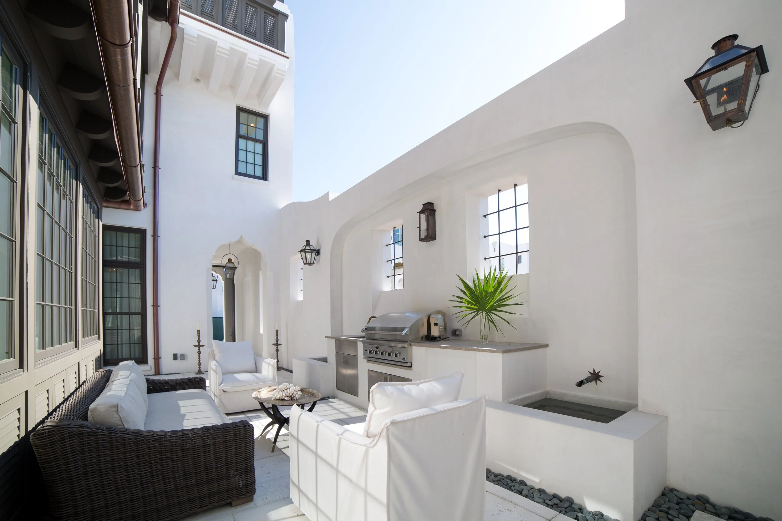 Outdoor patio with white walls, black lanterns, and windows; seating area with white cushions and a wicker sofa, a round table with coral decorations, a built-in grill, and a potted green plant