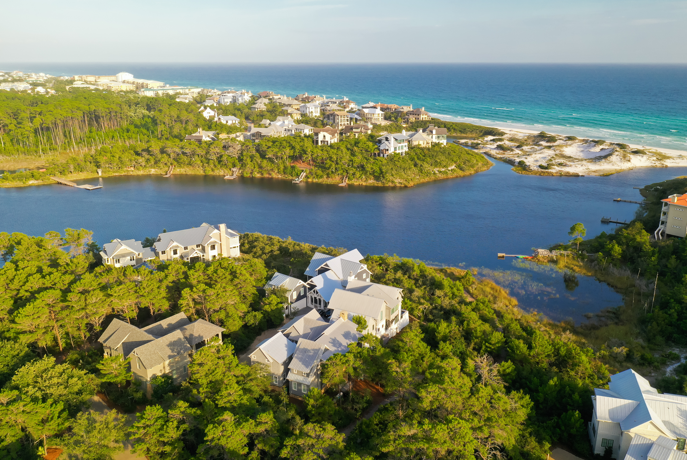 Aerial view of a coastal area showing houses surrounded by trees, a lagoon, and the ocean in the background.