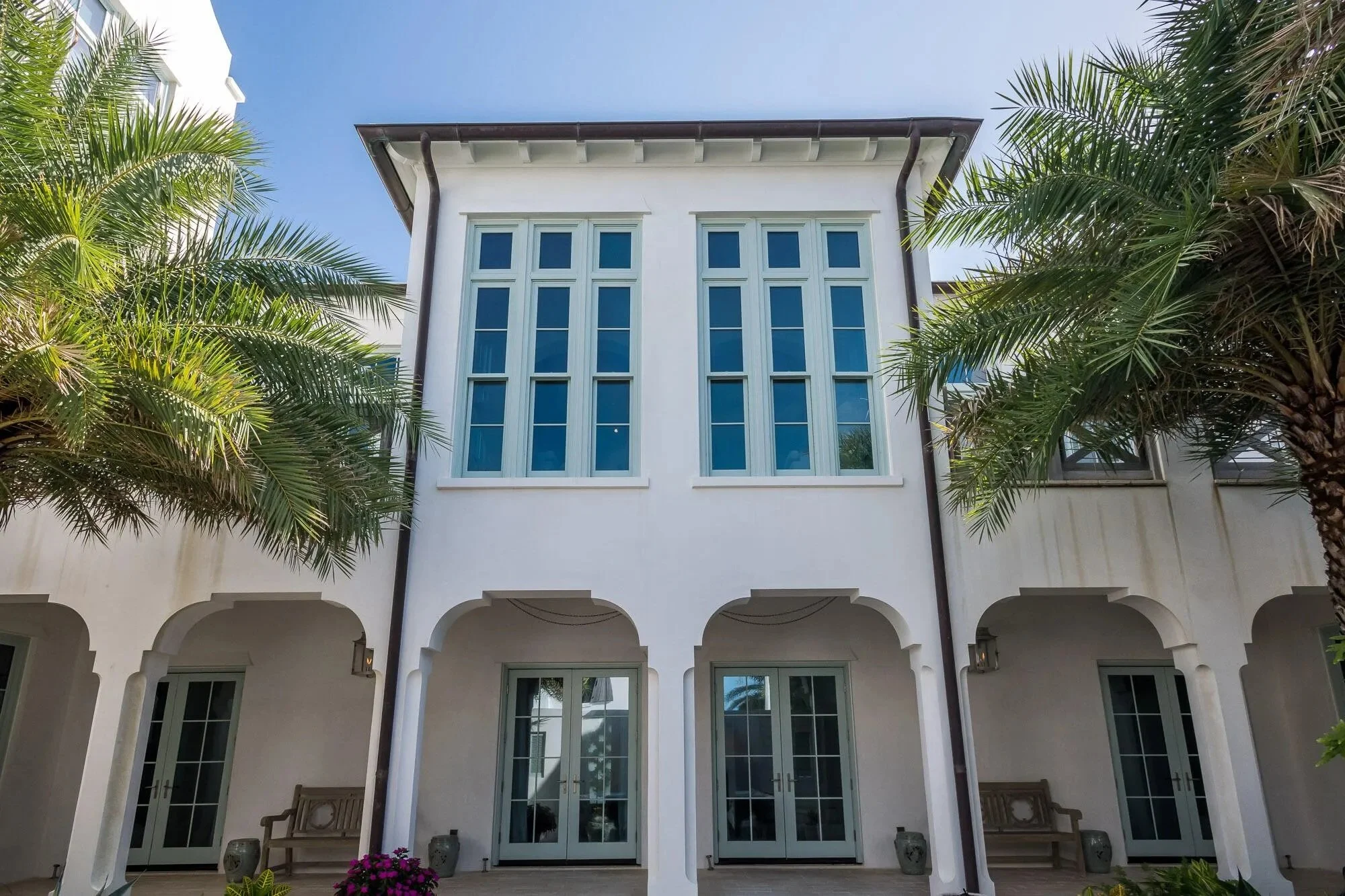 Front view of a white house with large windows, surrounded by palm trees, under a clear blue sky.