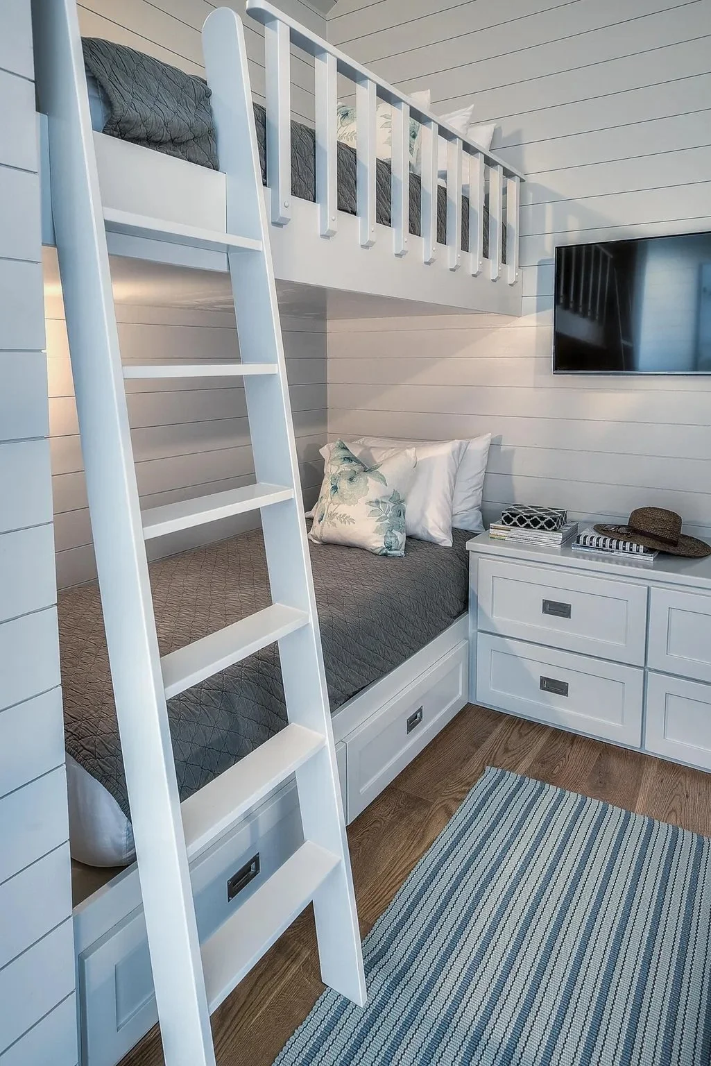 A white bunk bed with a ladder attached, set against a white paneled wall. The bottom bed has gray bedding and decorative pillows, with built-in drawers beneath. Beside the bed is a white dresser with drawers, atop which are books, a straw hat, and a