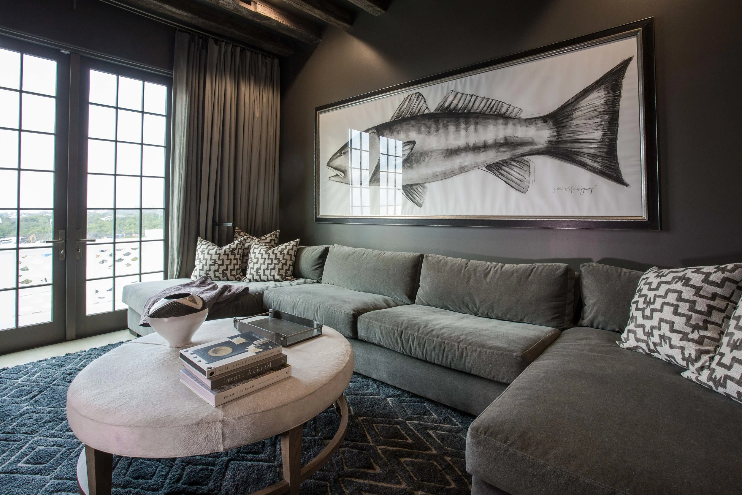 Living room with a large gray sectional sofa, decorative pillows, a round ottoman with books and a decorative object, dark wall, framed fish artwork, and glass doors with curtains.