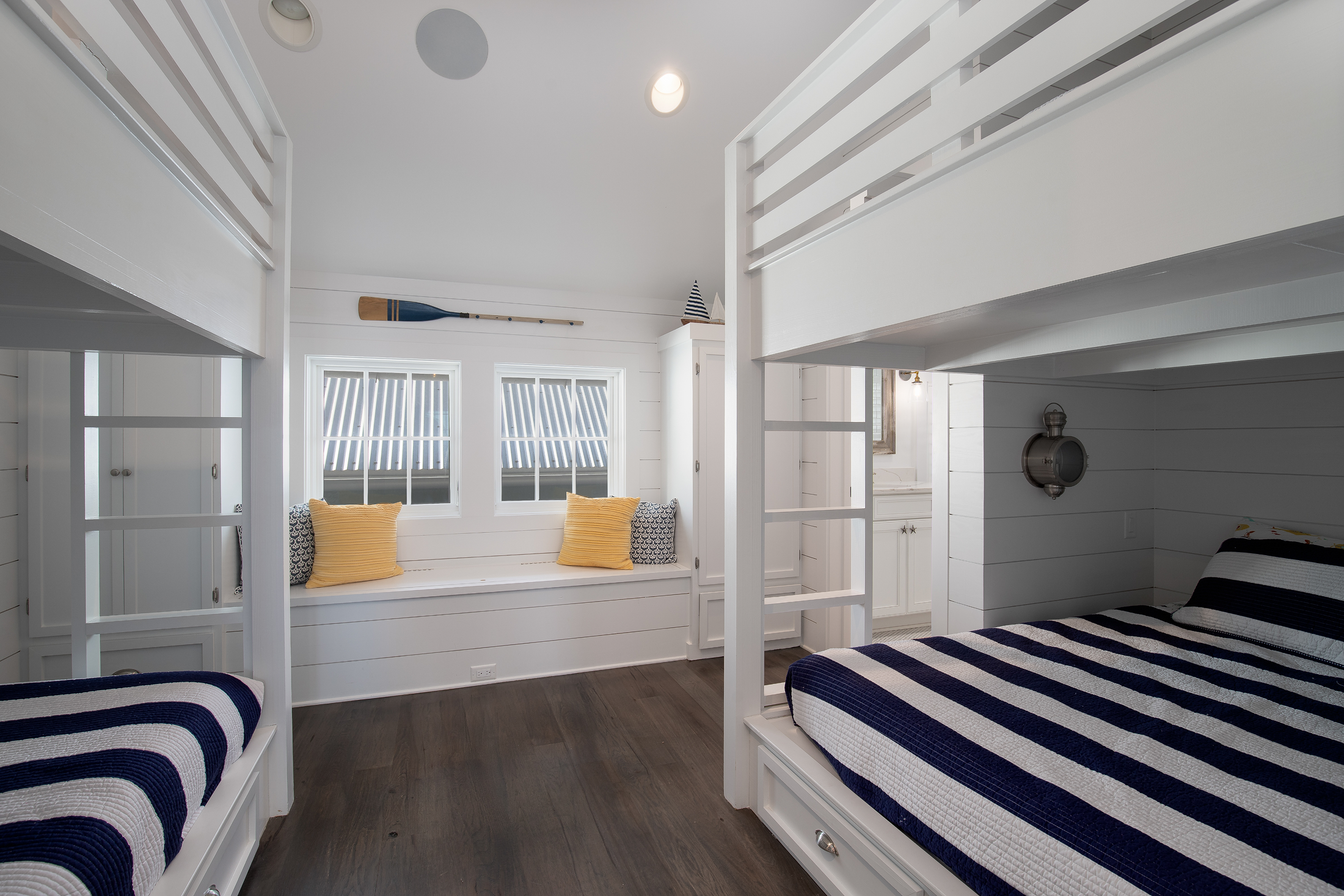Coastal-style bedroom with white wood paneling, bunk beds with striped bedding, a window seat with yellow and patterned pillows, and nautical decorations.