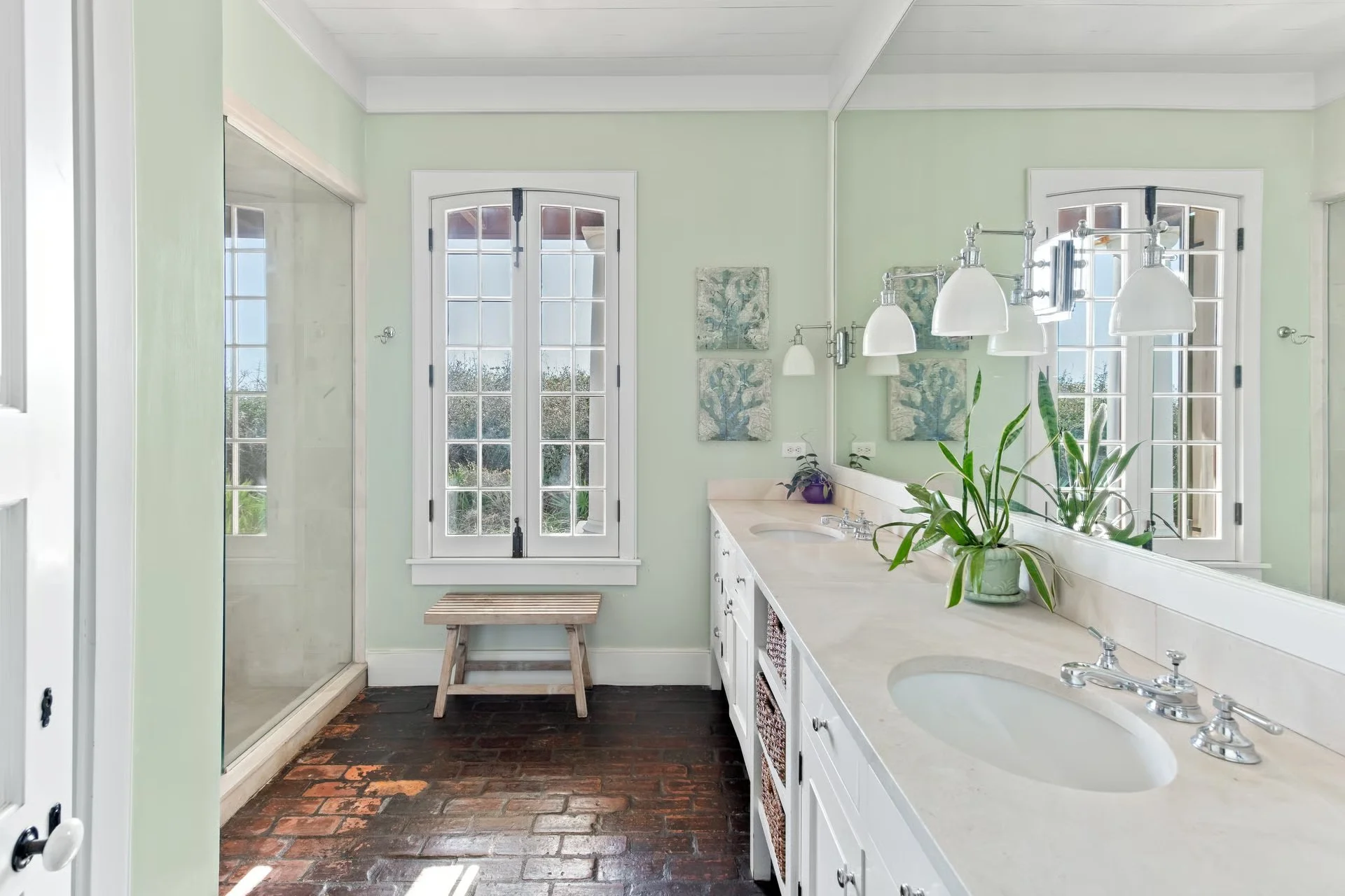 Bright bathroom with double vanity, large mirror, potted plant, framed artwork, and a shower with glass door, green walls, and brick floor.