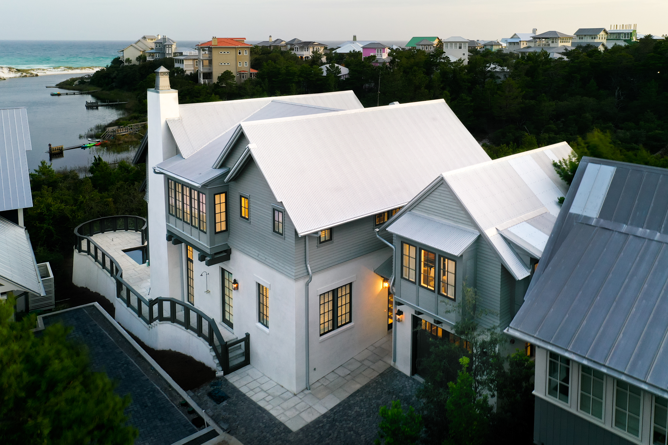 A large, modern house with a white and gray exterior, multiple sections with different roof heights, and illuminated windows overlooking a coastal landscape with other colorful houses and water.