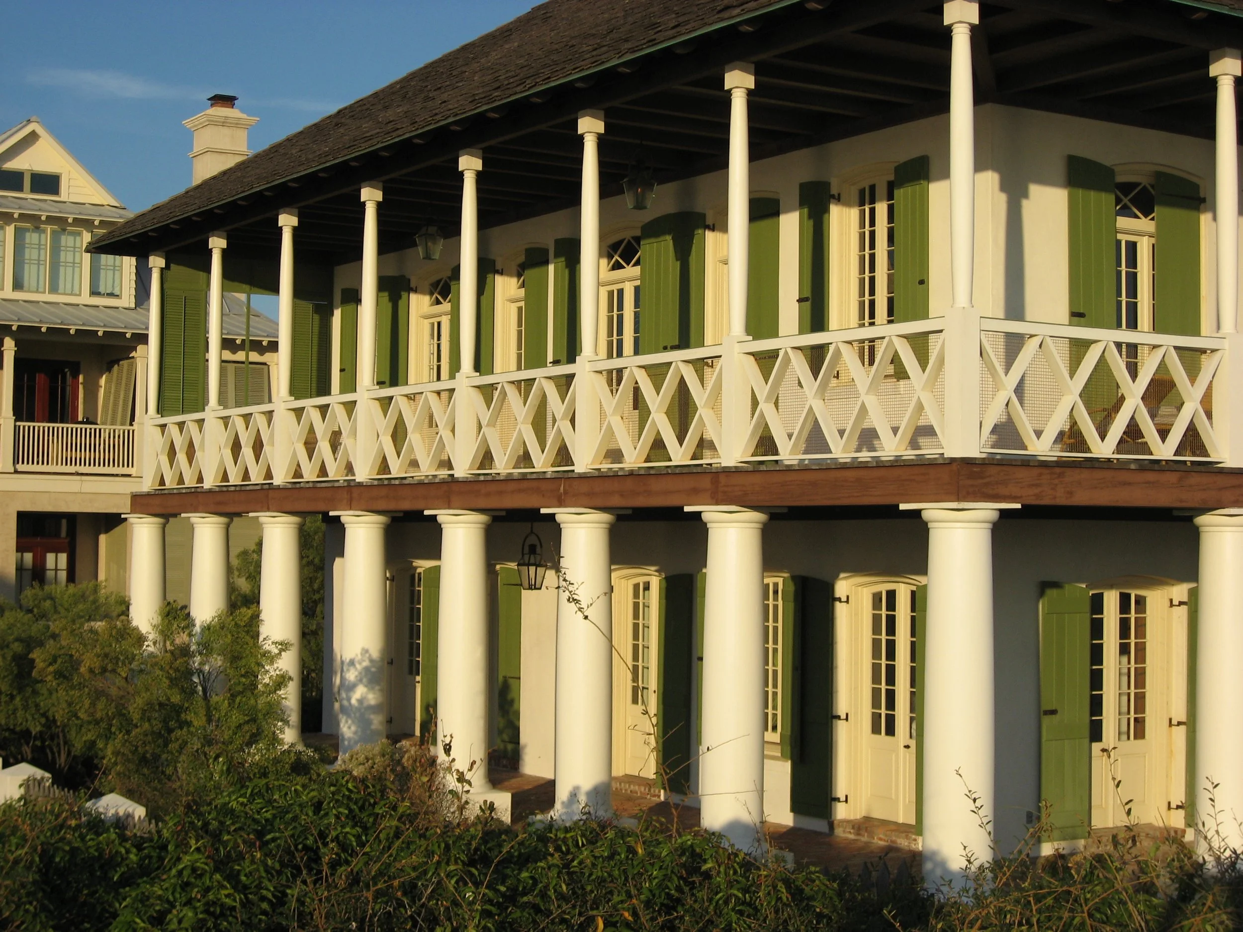 A two-story house with a balcony, green shutters, columns, and outdoor lighting, set against a clear blue sky.