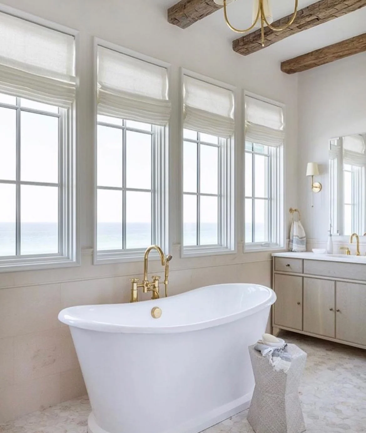 A bright bathroom with four large windows overlooking the ocean, a white bathtub with gold fixtures, a vanity with gold accents, and a small white textured stool with linens on top.
