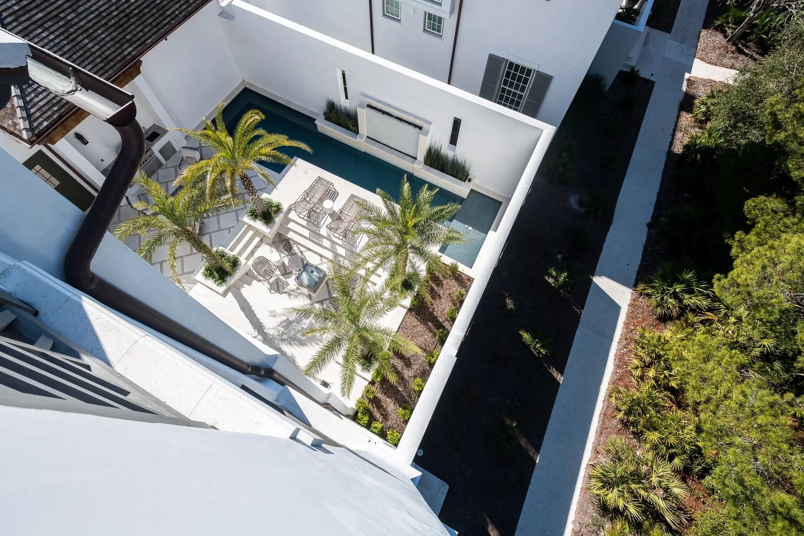 An aerial view of a modern apartment balcony with patio furniture, tall palm trees, a small swimming pool, and a landscaped yard with plants and trees.