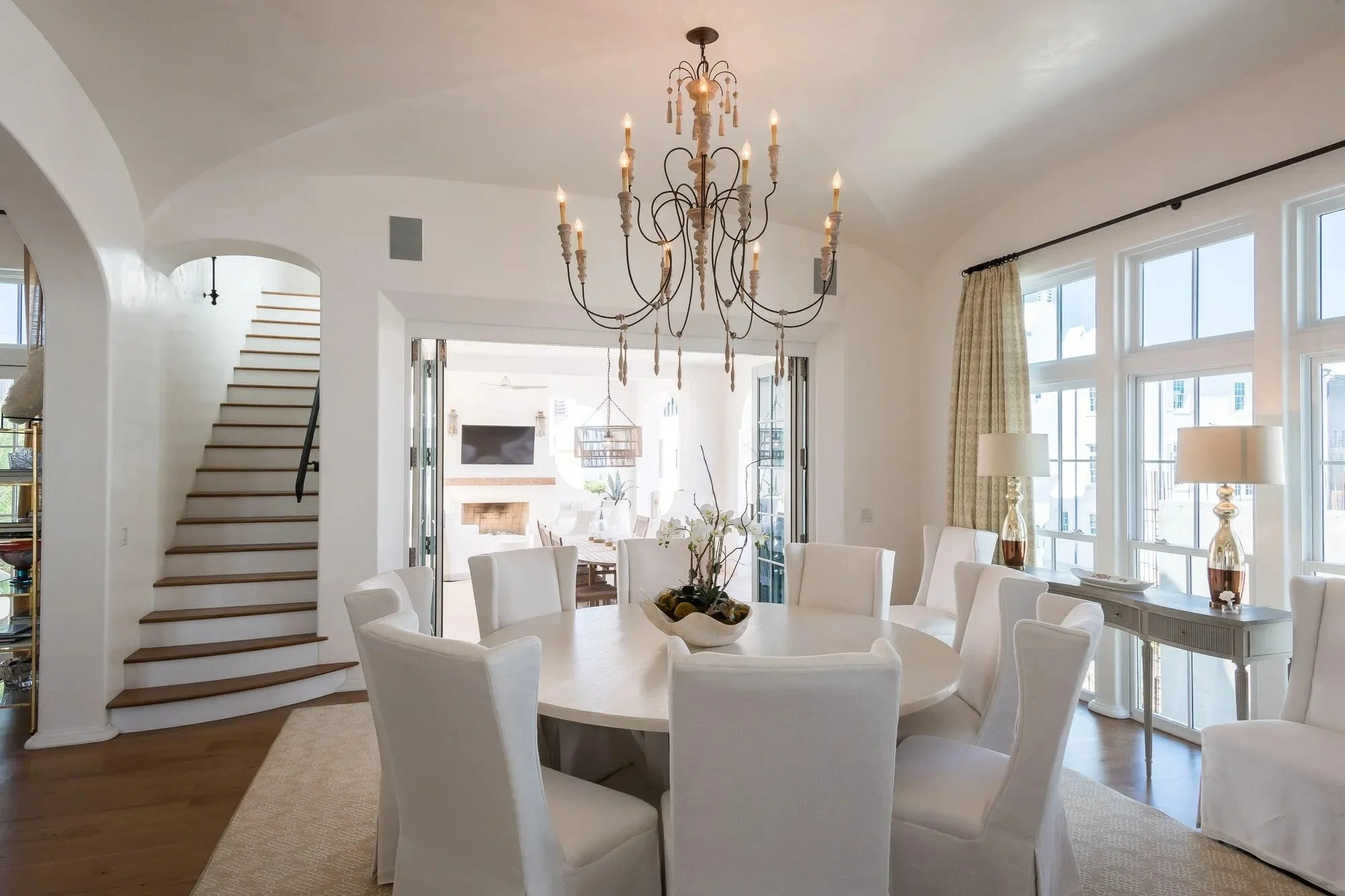 Bright, spacious dining room with a large round table surrounded by white upholstered chairs, a chandelier hanging overhead, large windows with curtains, and an adjoining living area