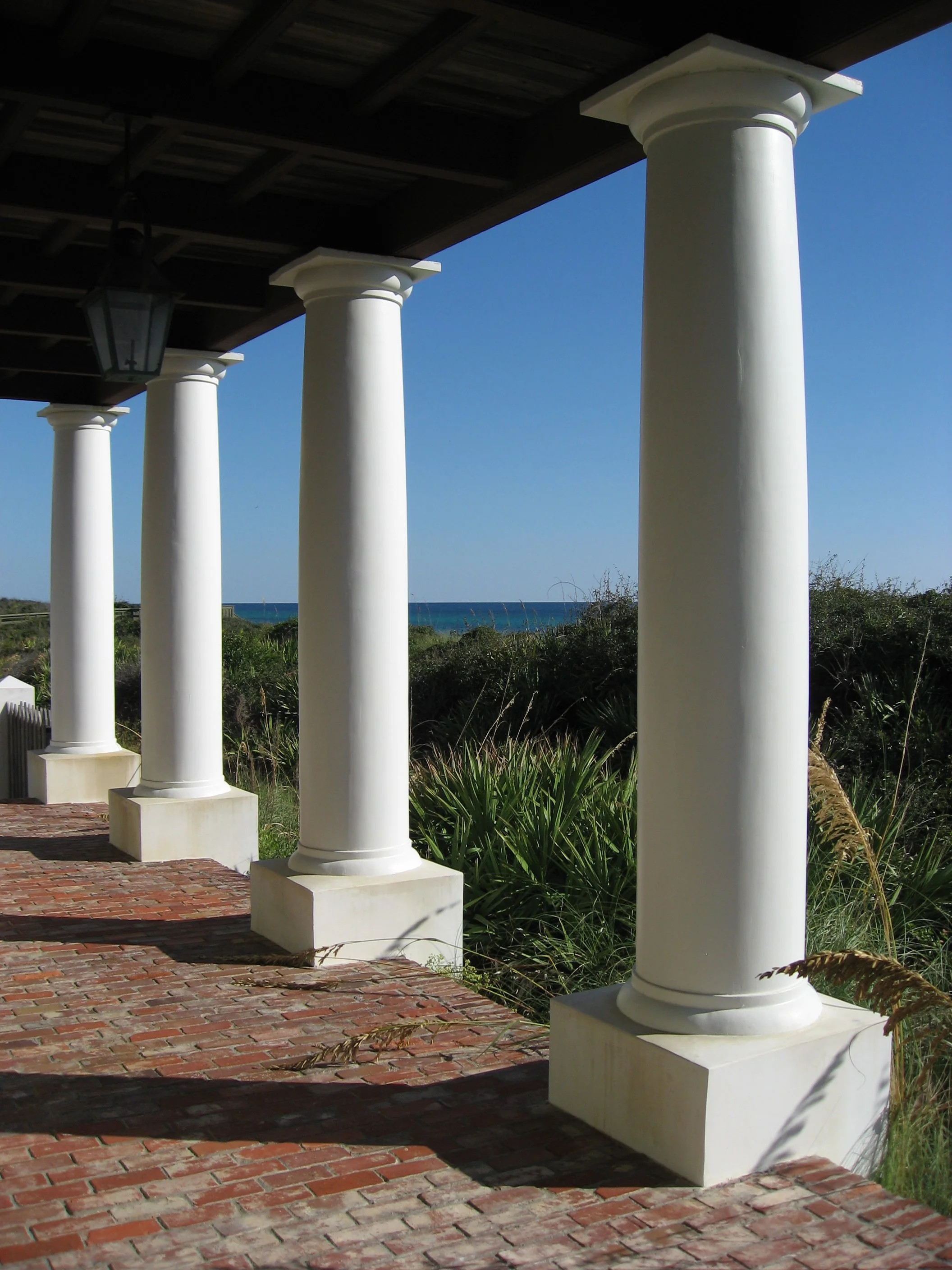 Four white columns supporting a roof over a brick-paved porch, with a view of the beach and ocean in the background.