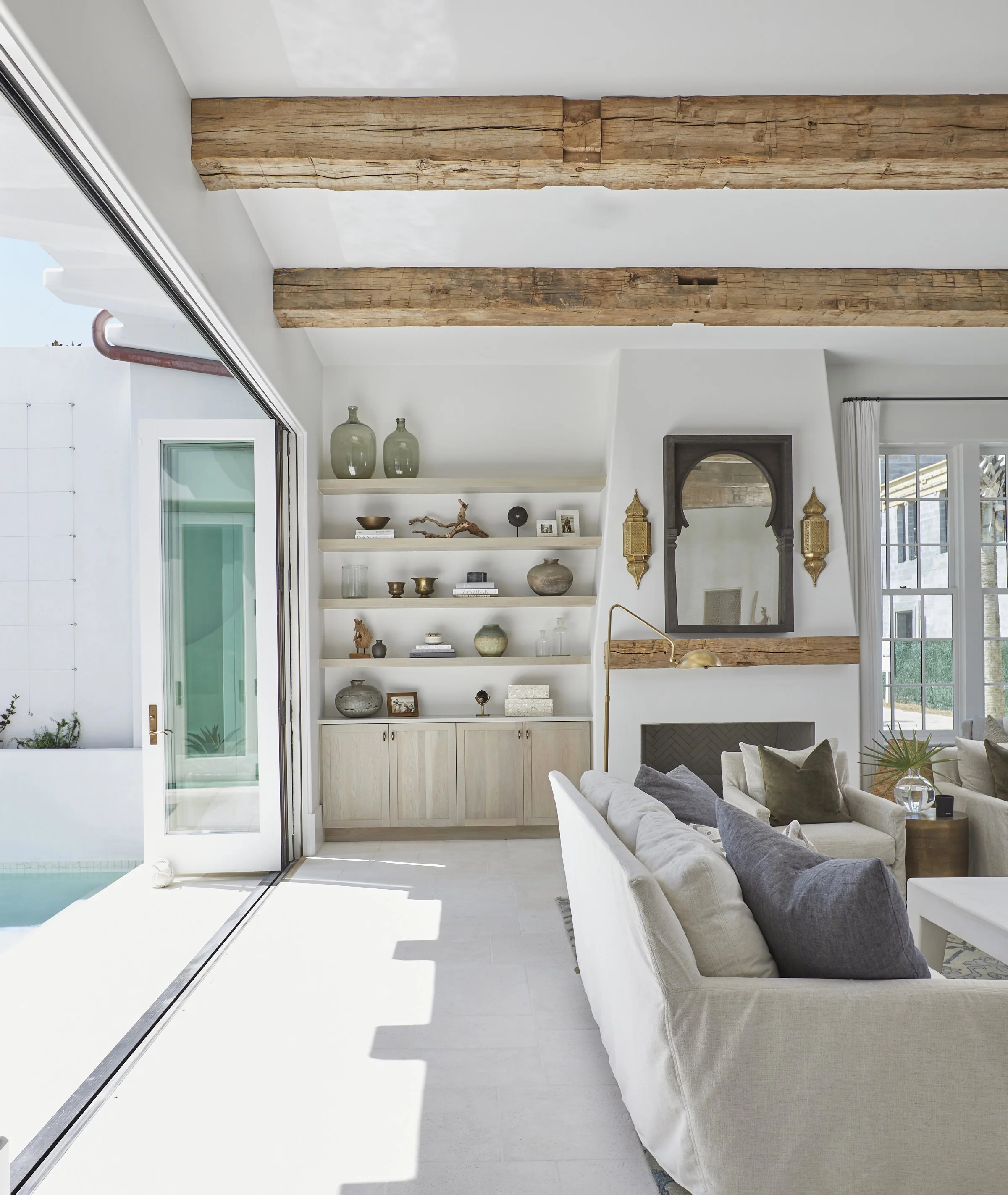 Modern living room with white walls, wooden ceiling beams, beige couch with dark pillows, and built-in shelves with decorative items. Large glass door opens to outdoor area.