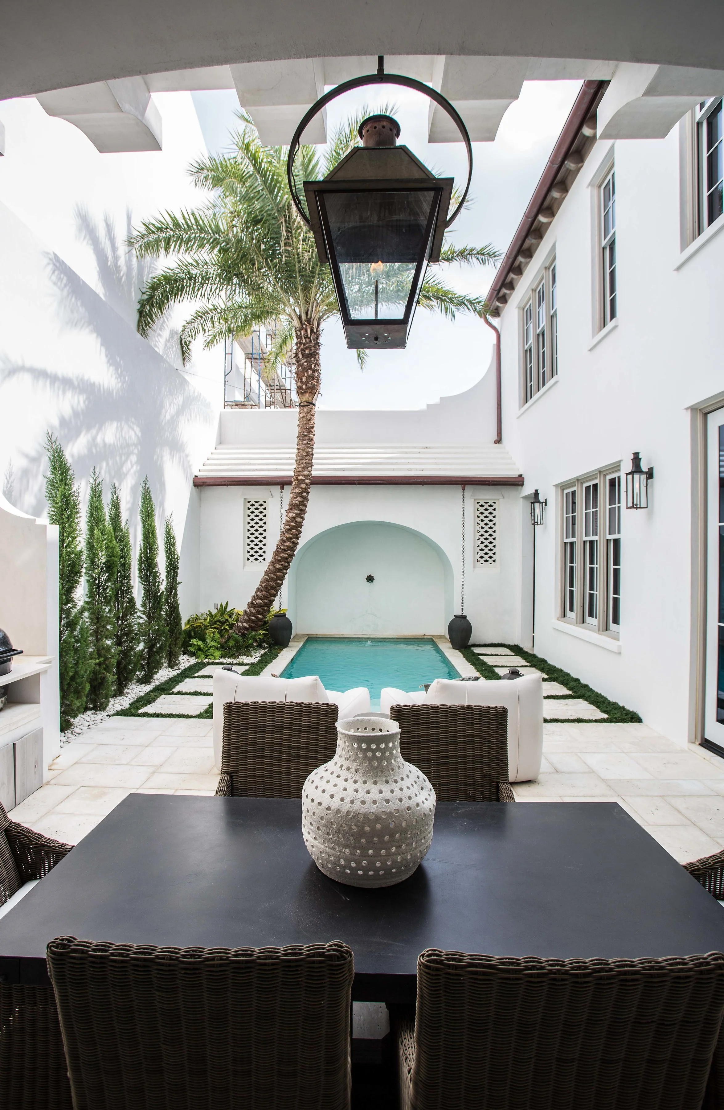Outdoor patio with dining table and chairs, overlooking a small swimming pool, palm tree, and white building walls, with lantern-style lights.