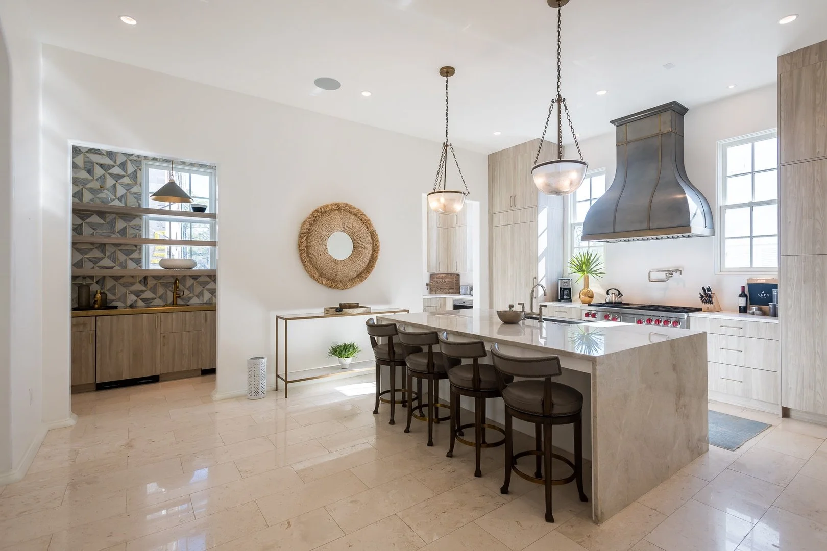 Modern kitchen with white walls, a large island with a marble countertop, five brown chairs, and pendant lighting. Contains a stainless steel range hood, light wood cabinetry, and a window with natural light.