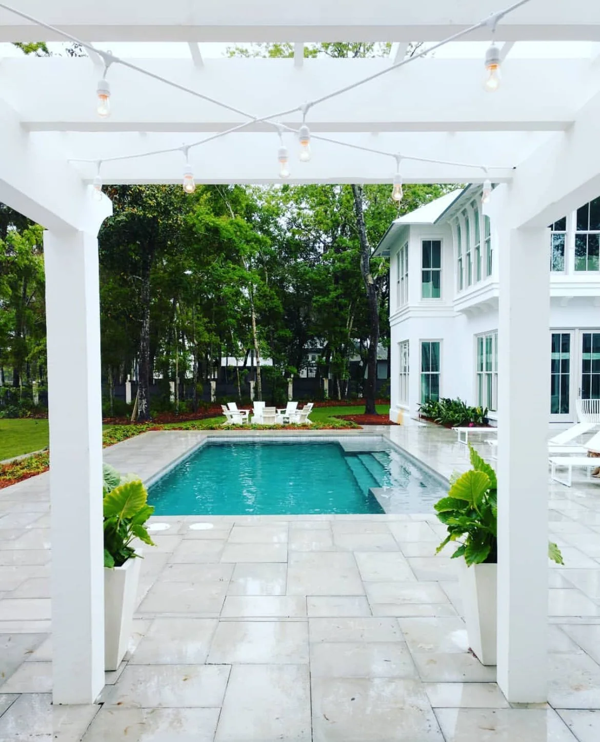 View of a backyard patio with a swimming pool, white chairs, lush green trees, and a white house in the background.