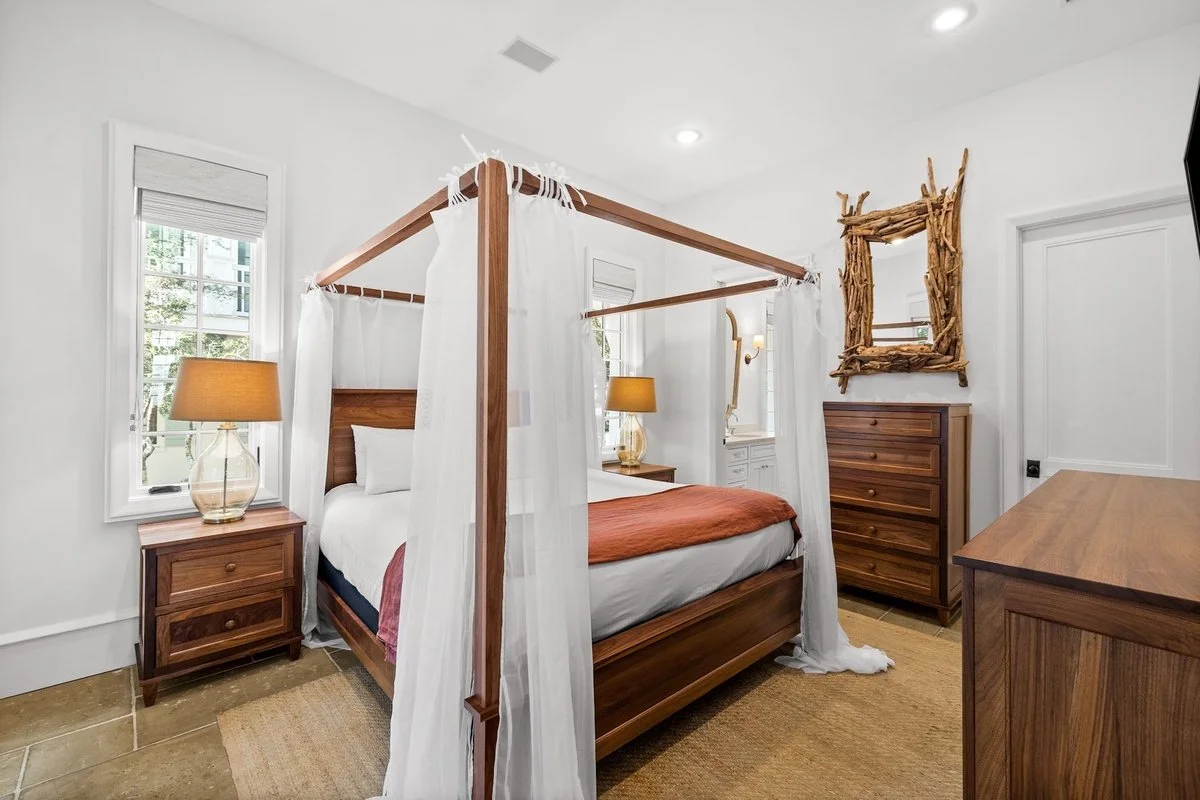 A bedroom with a four-poster bed decorated with white curtains and a brownish-orange bedspread. There are two windows with white blinds, a wooden bedside table with a glass lamp, and a wooden dresser. A rustic mirror made of driftwood hangs on the wa