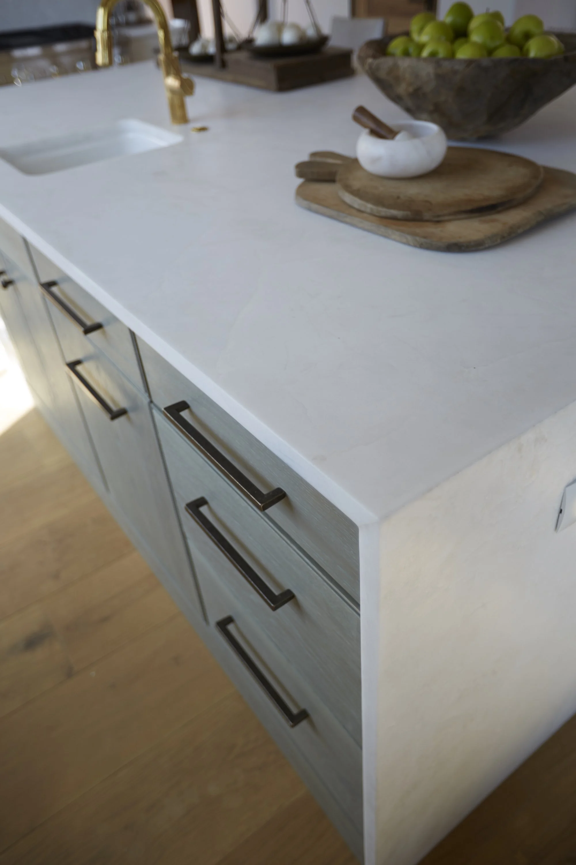 Close-up of a kitchen countertop with a white surface, featuring a sink, a gold faucet, a wooden cutting board, a white mortar with a pestle, and a large wooden bowl filled with green apples.