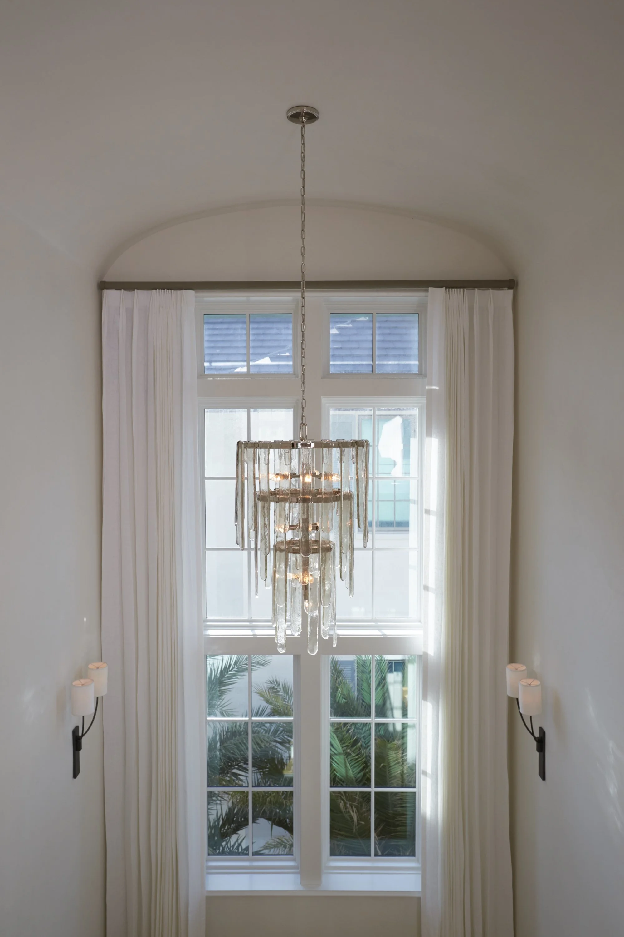 Interior view of a room with large double-height windows, white curtains, a chandelier hanging from the ceiling, and two wall-mounted sconces on each side of the windows. Outside, palm trees are visible through the windows.