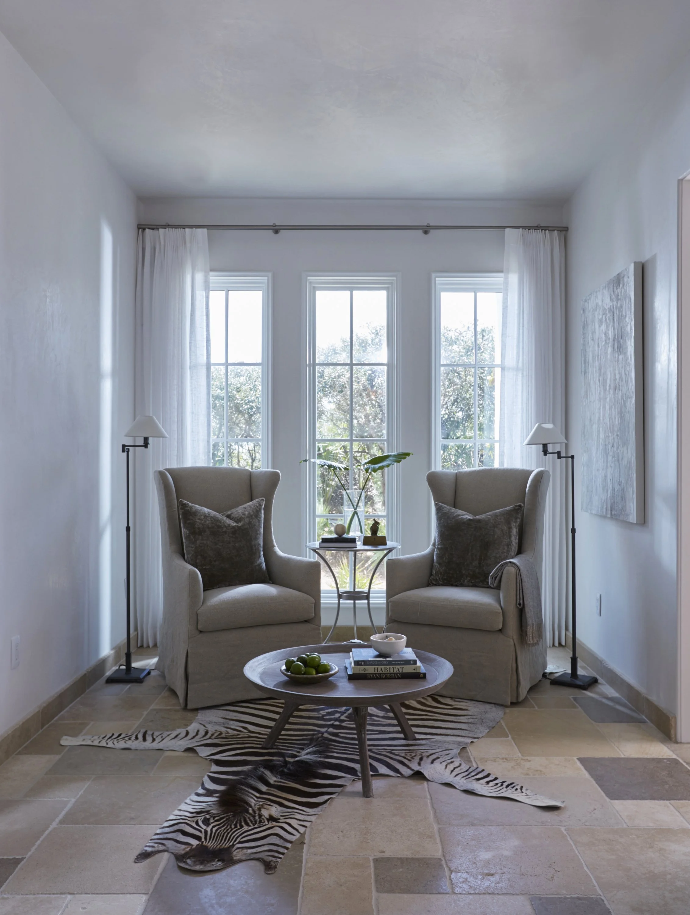 Living room with two armchairs, a small round table, a zebra-print rug, and large windows letting in natural light.