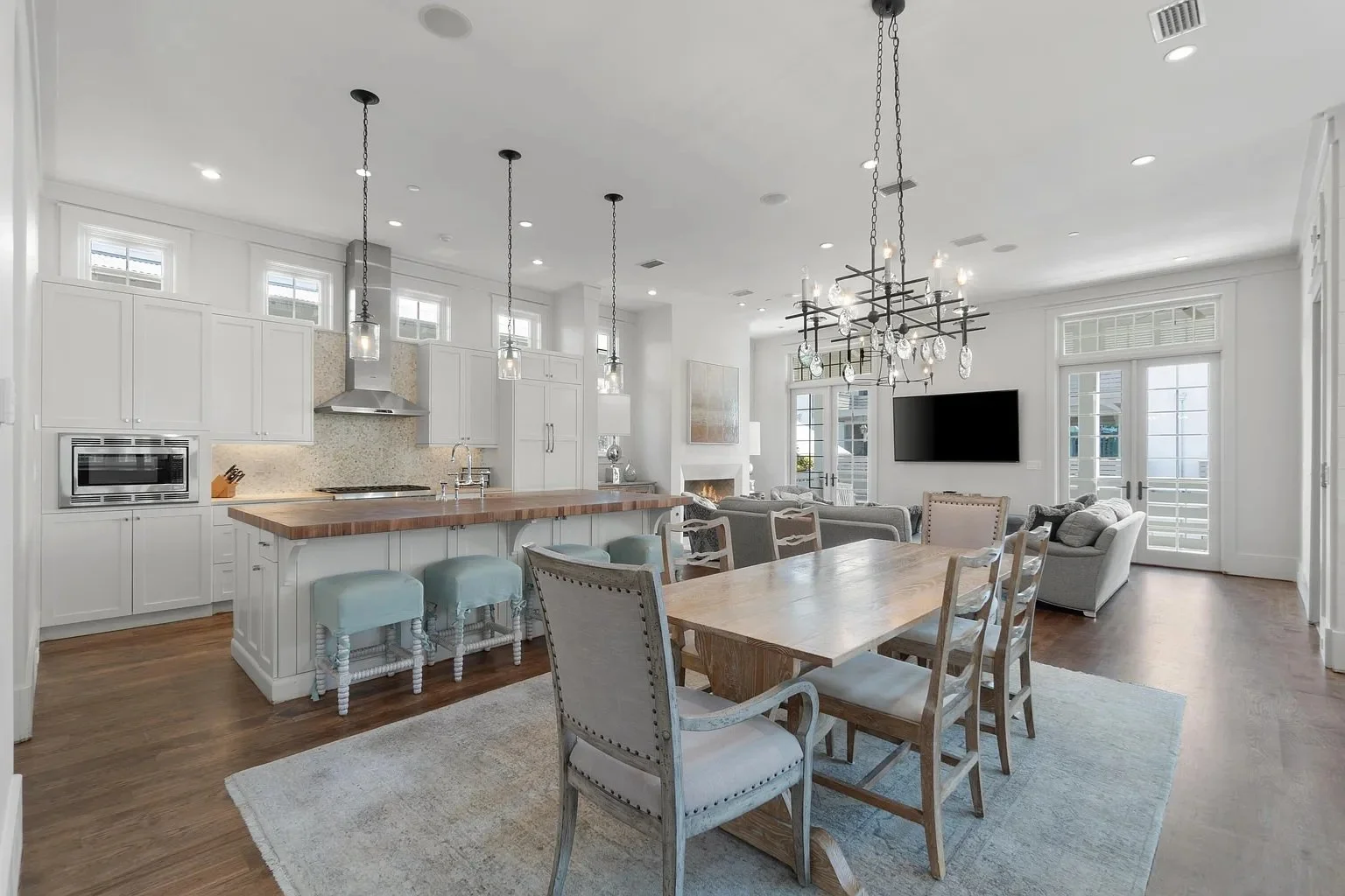Open-concept living room and kitchen with white cabinetry, wood accents, and modern decor, including a dining table with mixed chairs, a chandelier, and multiple windows letting in natural light.