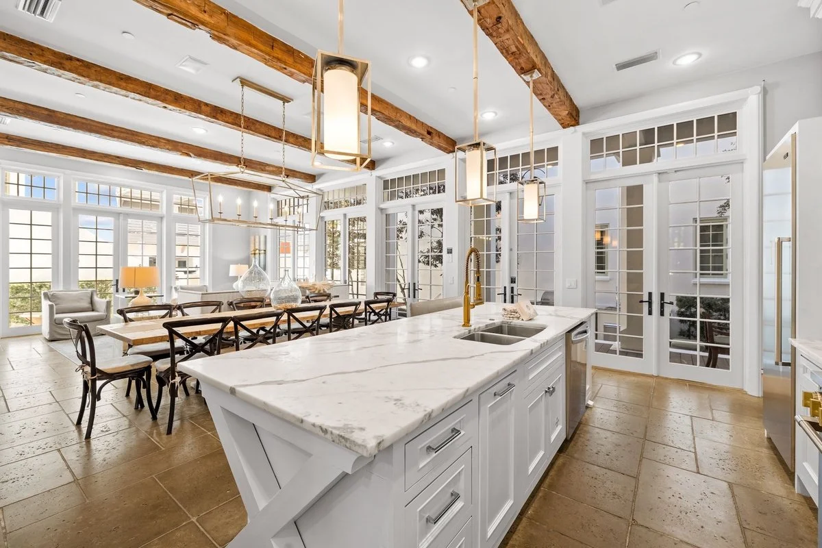 A bright open-concept kitchen and dining area with large windows, white cabinets, a marble island with a gold faucet, wooden beams on the ceiling, and a long dining table with black chairs.