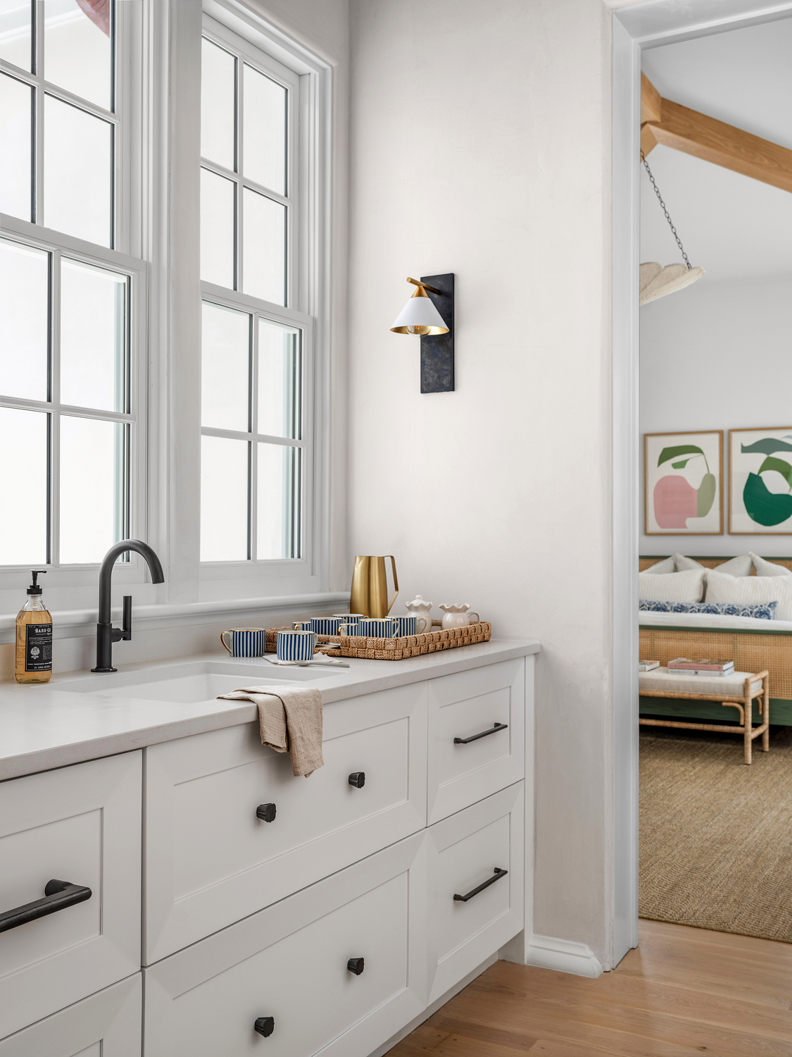 A kitchen with white cabinetry and a white countertop, black hardware, and a black faucet. On the counter, there is a soap dispenser, a tray with blue and white striped cups, and a gold pitcher. There are large windows above the sink, and a towel han