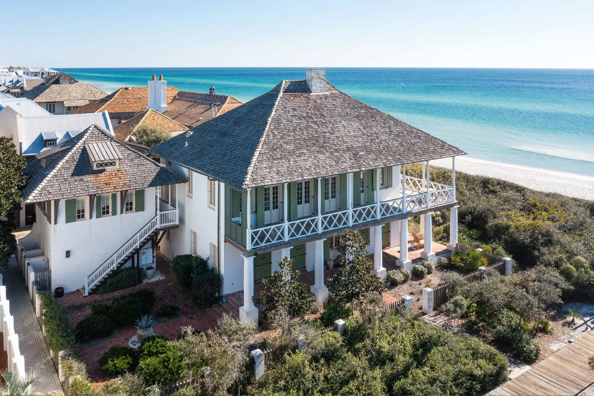 A large beach house with a wooden shingle roof and a wraparound porch, overlooking the ocean with turquoise waves and a sandy beach. Nearby houses and lush greenery surround the property.