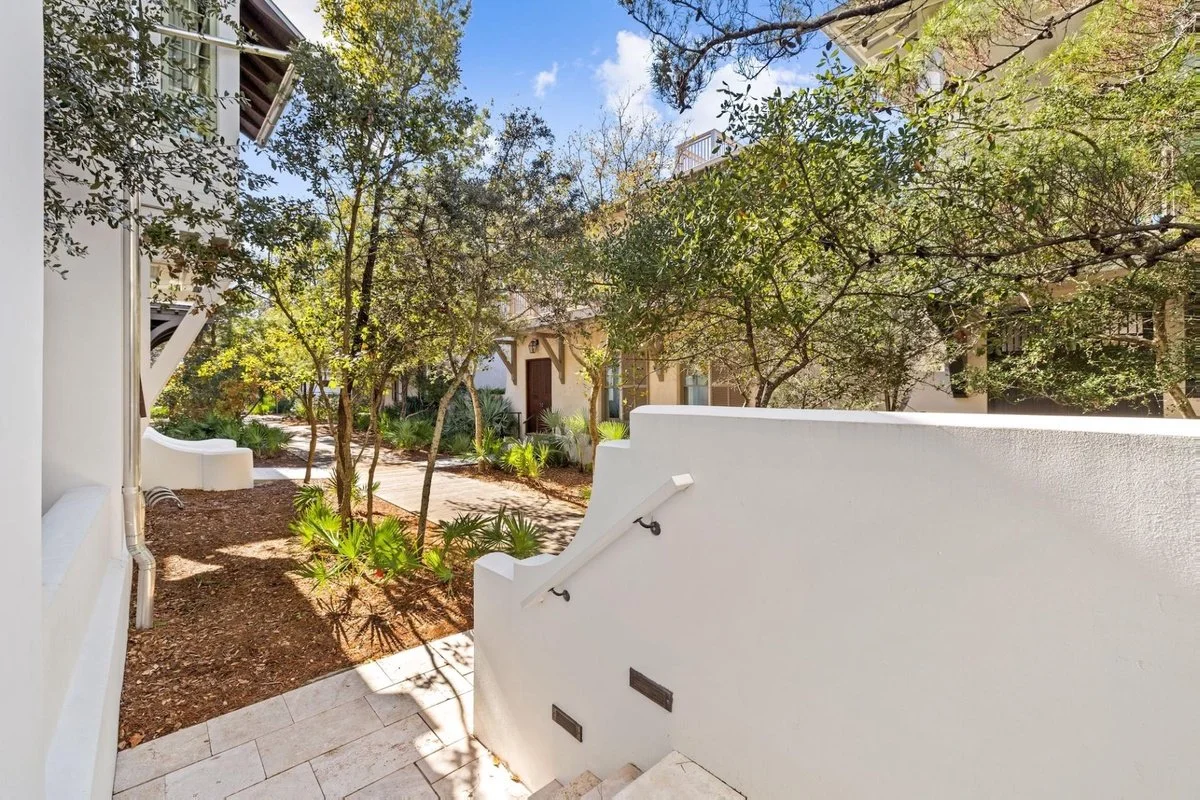 A small outdoor courtyard with white stucco walls, trees, and plants, and a walkway with stairs leading down, in a sunny setting.