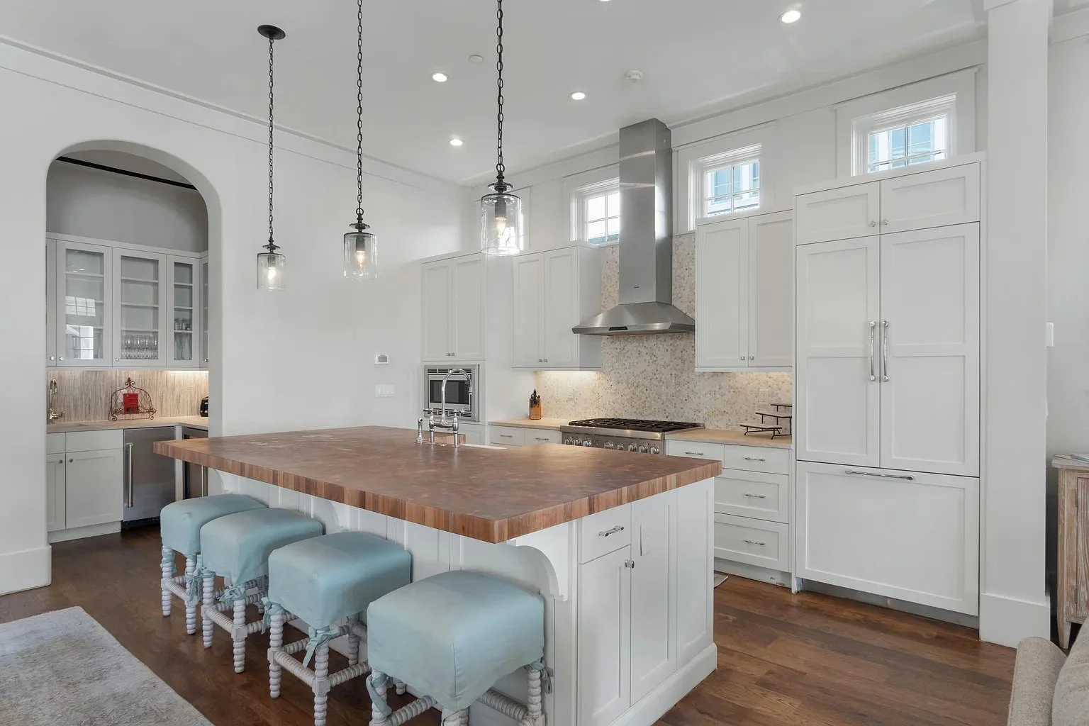 Modern white kitchen with wooden island counter, three light fixtures hanging above, stainless steel stove and hood, built-in microwave, and cabinets with farmhouse style.