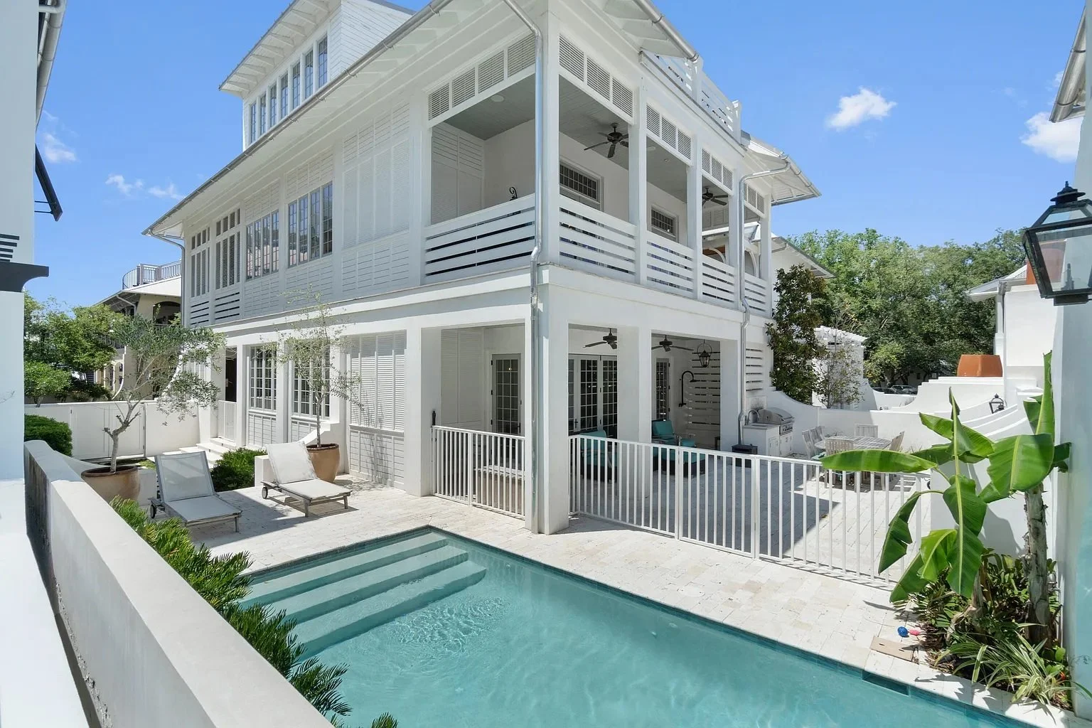 White multi-story house with screened porches, a small backyard pool, potted plants, lounge chairs, and outdoor seating under a bright blue sky.
