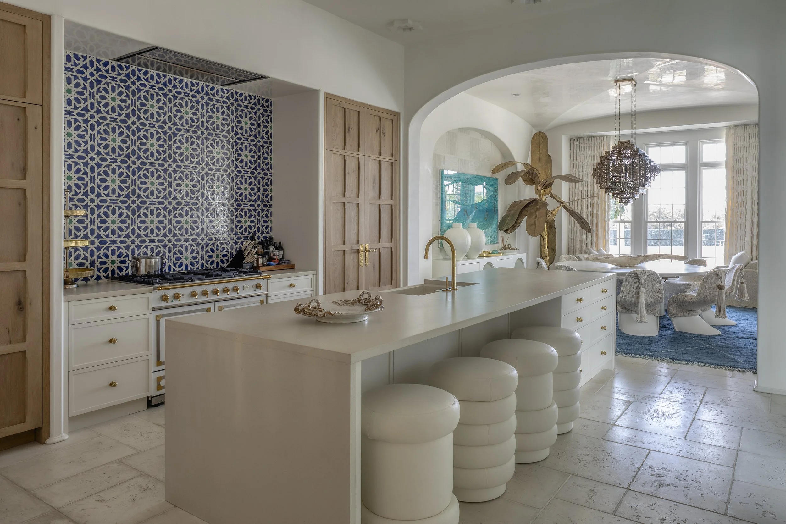 Modern kitchen and dining area with a white island, bar stools, a stove, and a colorful blue patterned backsplash, leading to a cozy dining space with a round table, chairs, large windows, and elegant light fixtures.