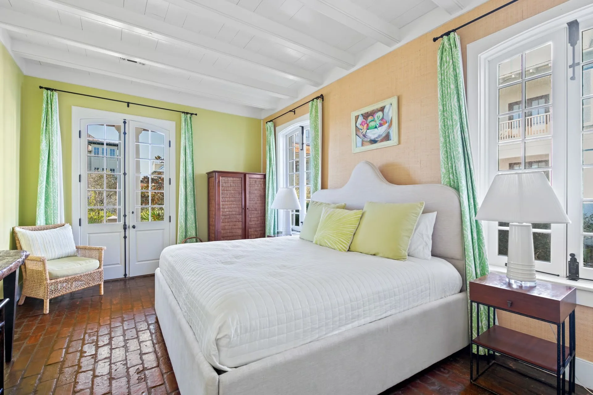 A bright bedroom with yellow and beige walls, white wooden ceiling, and large windows with green curtains. There's a white bed with yellow pillows, a wooden bedside table with a white lamp, a wicker chair with a striped pillow, and a wooden cabinet.