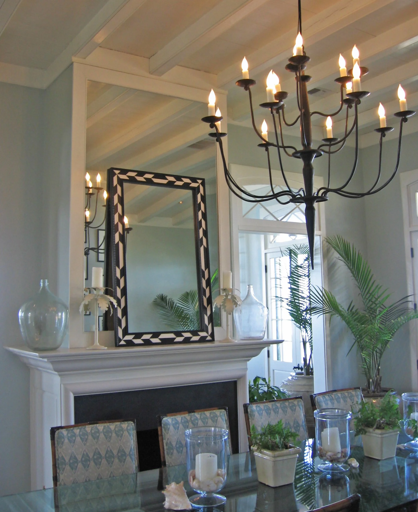 A dining room with a black chandelier with candle-shaped lights, a mirror above the fireplace, and potted plants near the window.