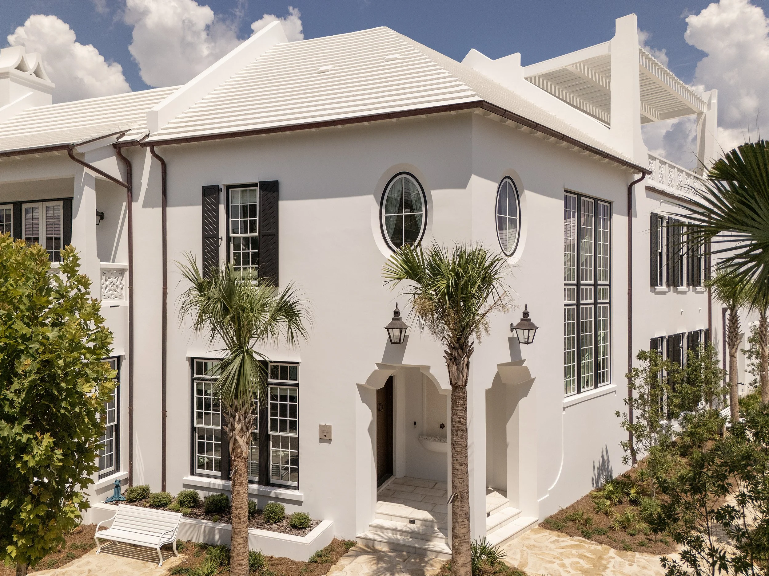 White house with black shutters, palm trees, and a small outdoor sitting area under a partly cloudy sky.