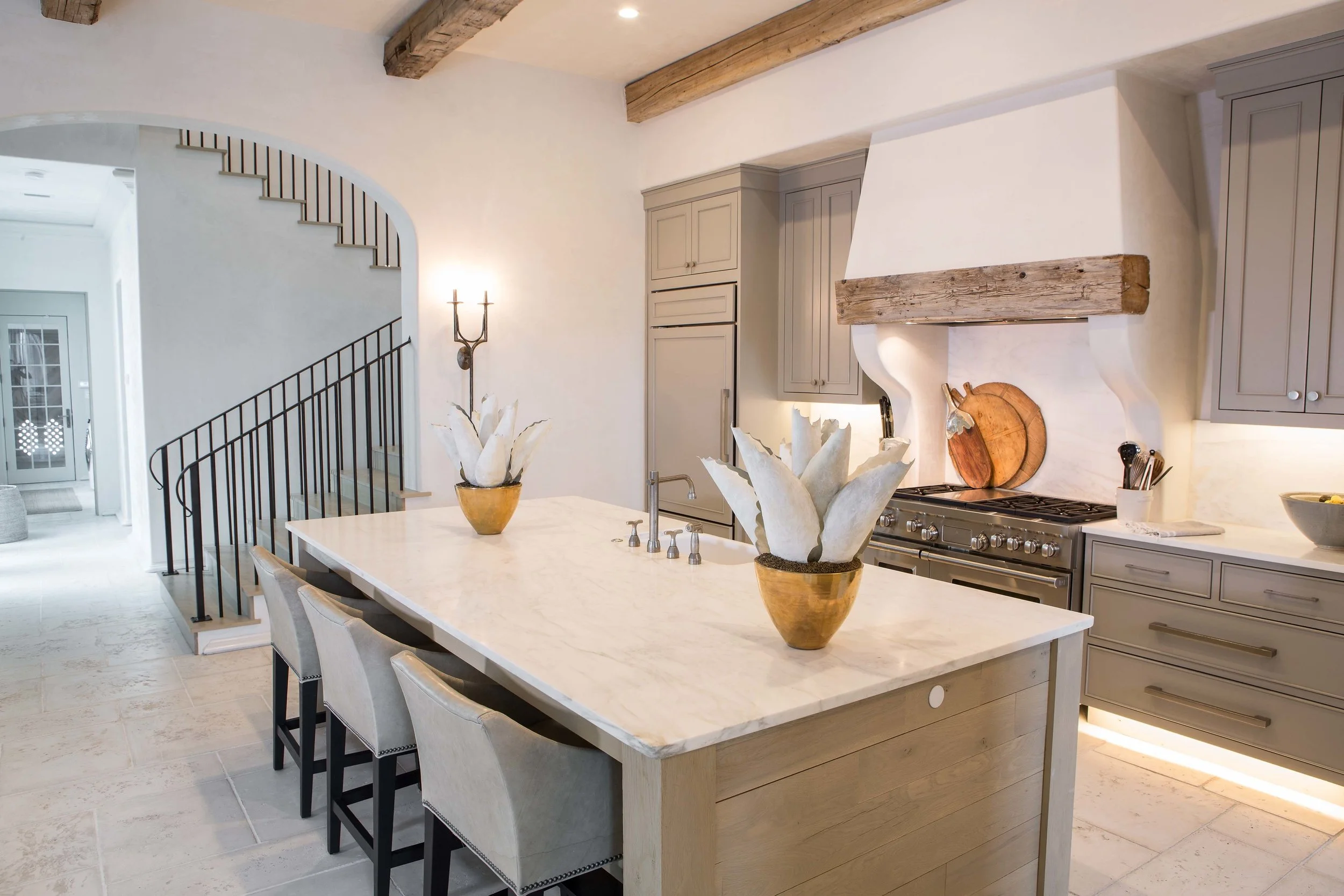 Modern kitchen with a white marble island, light gray cabinetry, and wooden beams on the ceiling. Decor includes potted plants on the island and a large cutting board near the stove.