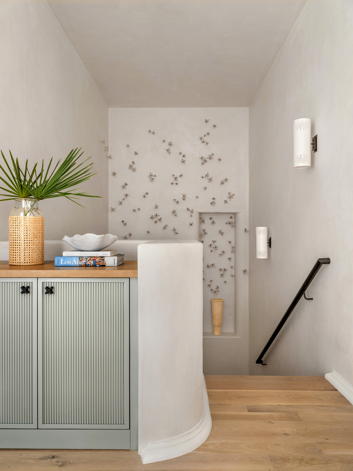 Interior view of a corner with a staircase, a light green cabinet, a potted plant, books, and decorative wall art.