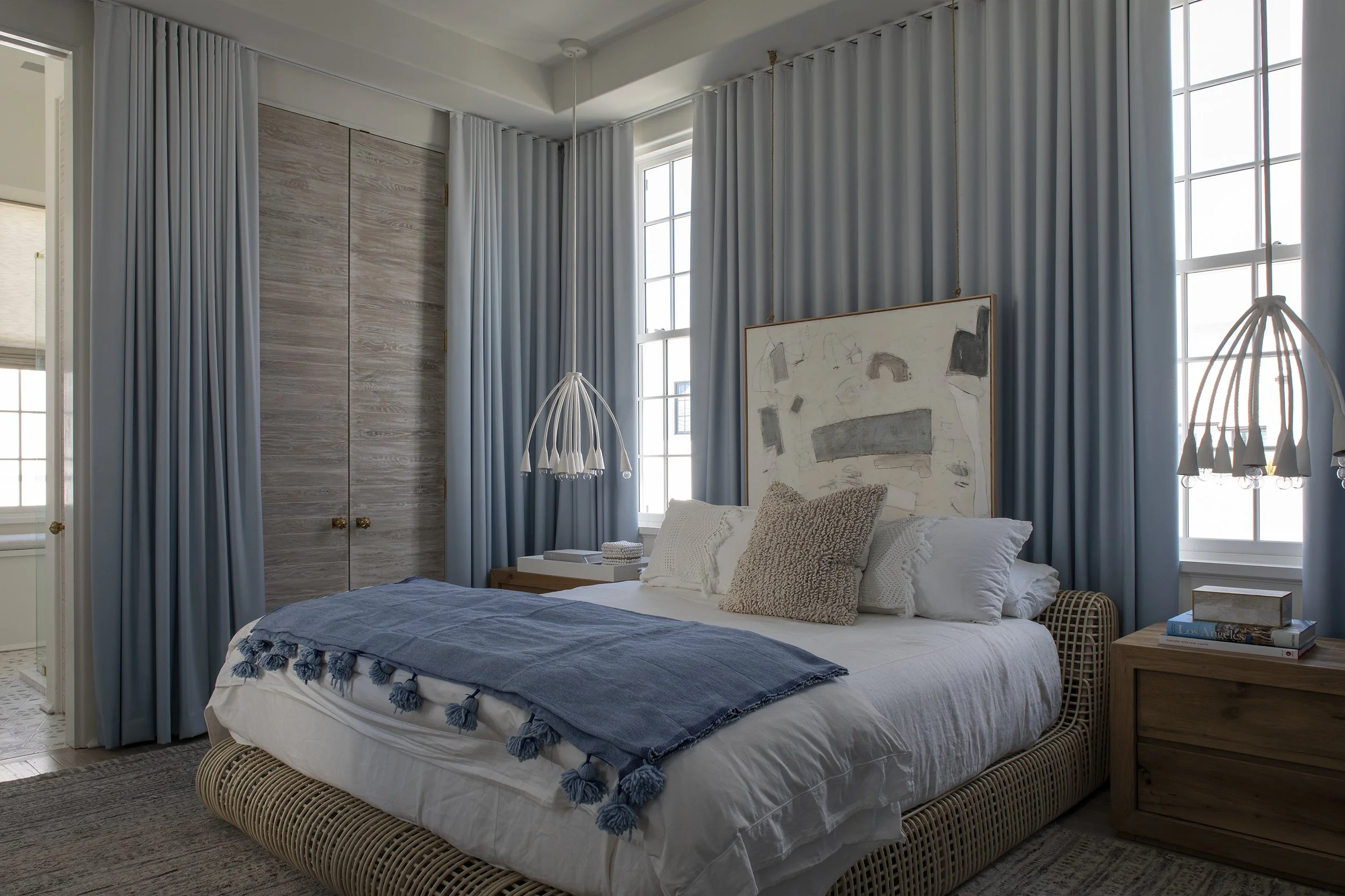 A cozy bedroom with a bed, blue and beige accents, a nightstand with books, and large windows with blue curtains.