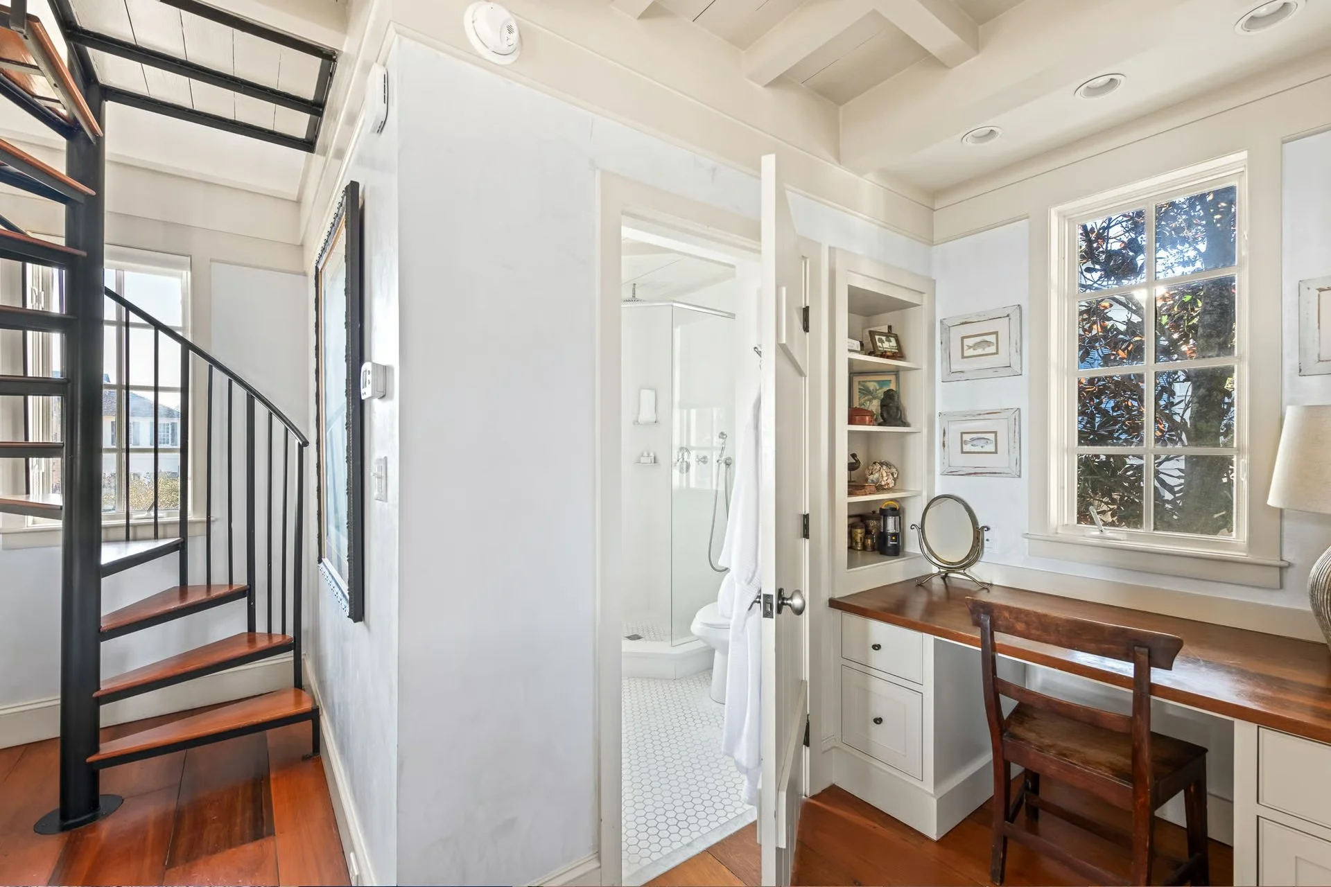 Interior view of a small room with a bathroom door open, showing a shower, a window with trees outside, and a built-in wooden desk with a chair. There are picture frames on the wall and a small shelf with decorative items. A spiral staircase is visib