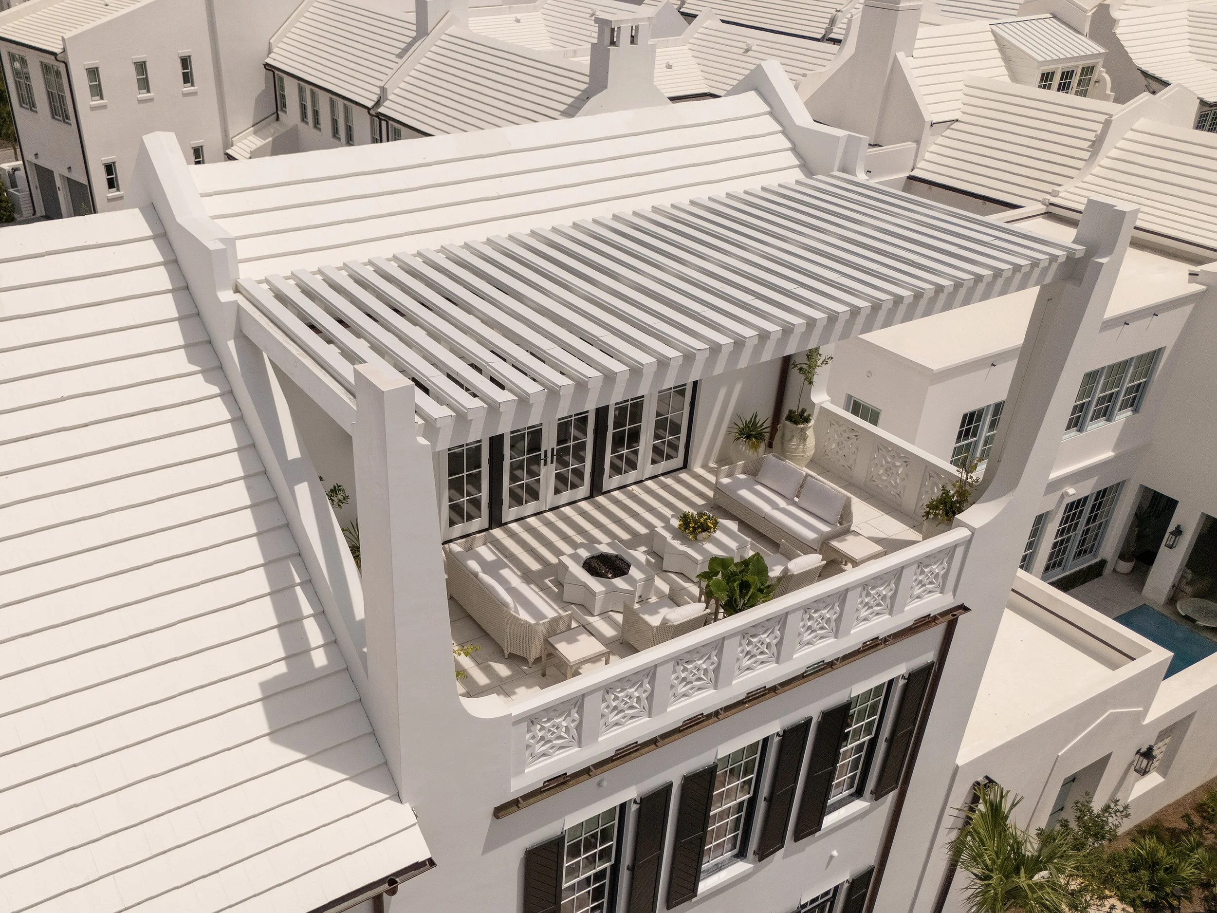 A white rooftop terrace with outdoor furniture, potted plants, and a pergola, surrounded by white houses with similar rooftops.