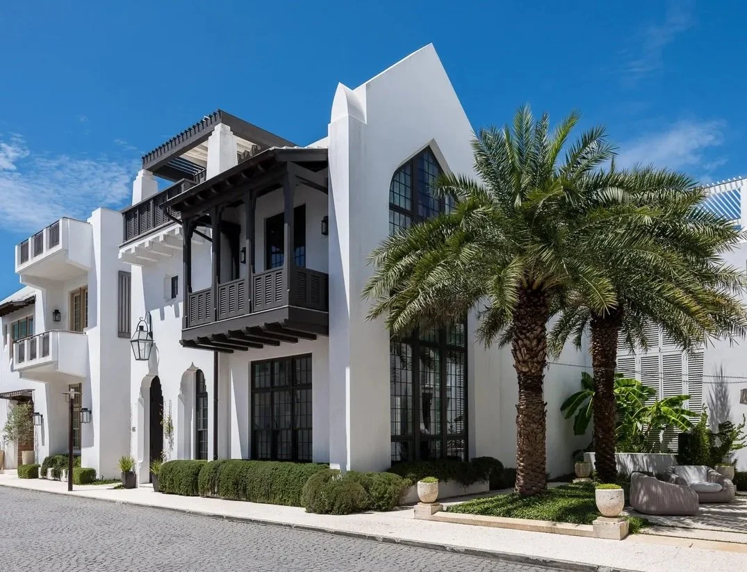 White modern building with black accents, large windows, and balconies, surrounded by palm trees on a sunny day.