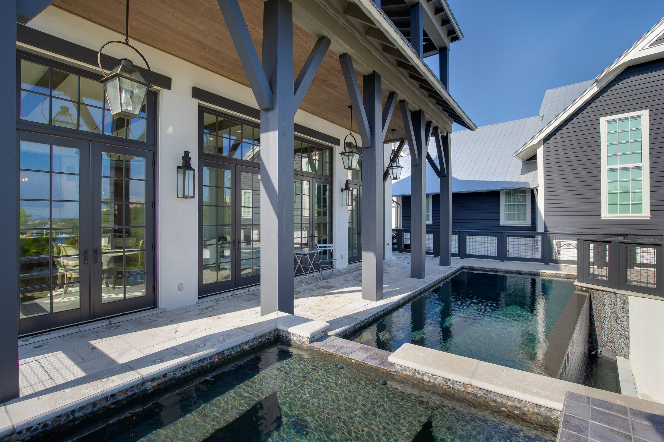 A backyard patio area with a small pool, glass double doors, black exterior walls, and black support beams under a wooden ceiling with hanging lanterns.