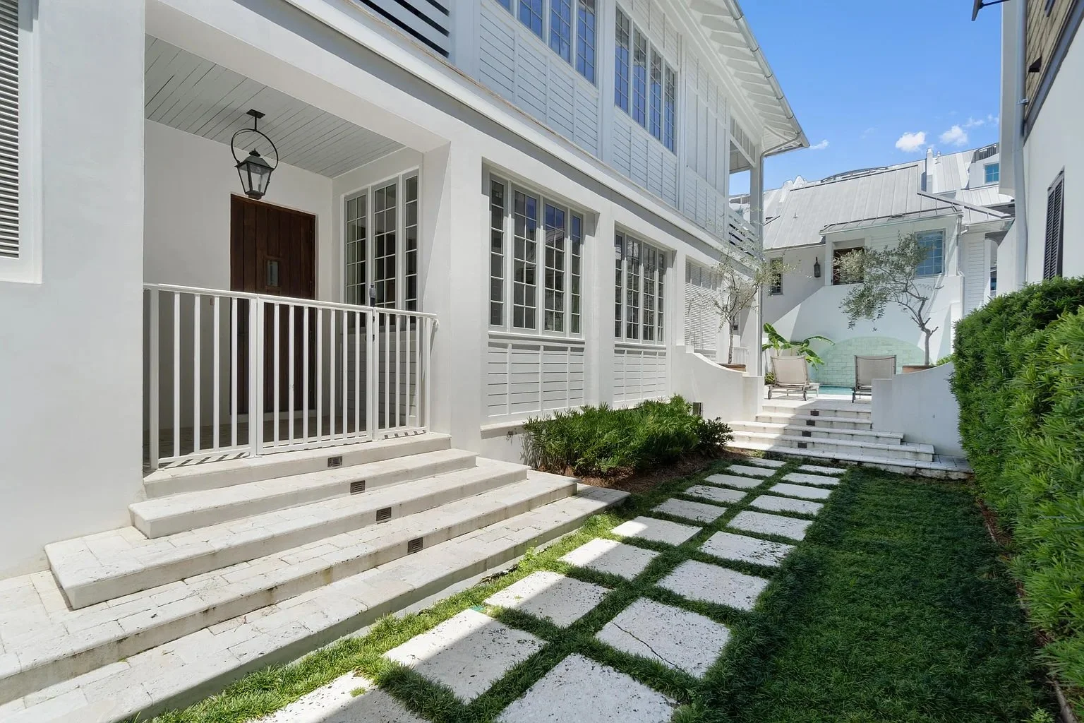 Exterior view of a modern white house with a porch, steps leading up to the front door, a gated entrance, and a landscaped yard with stepping stones, bushes, and trees under a bright blue sky.