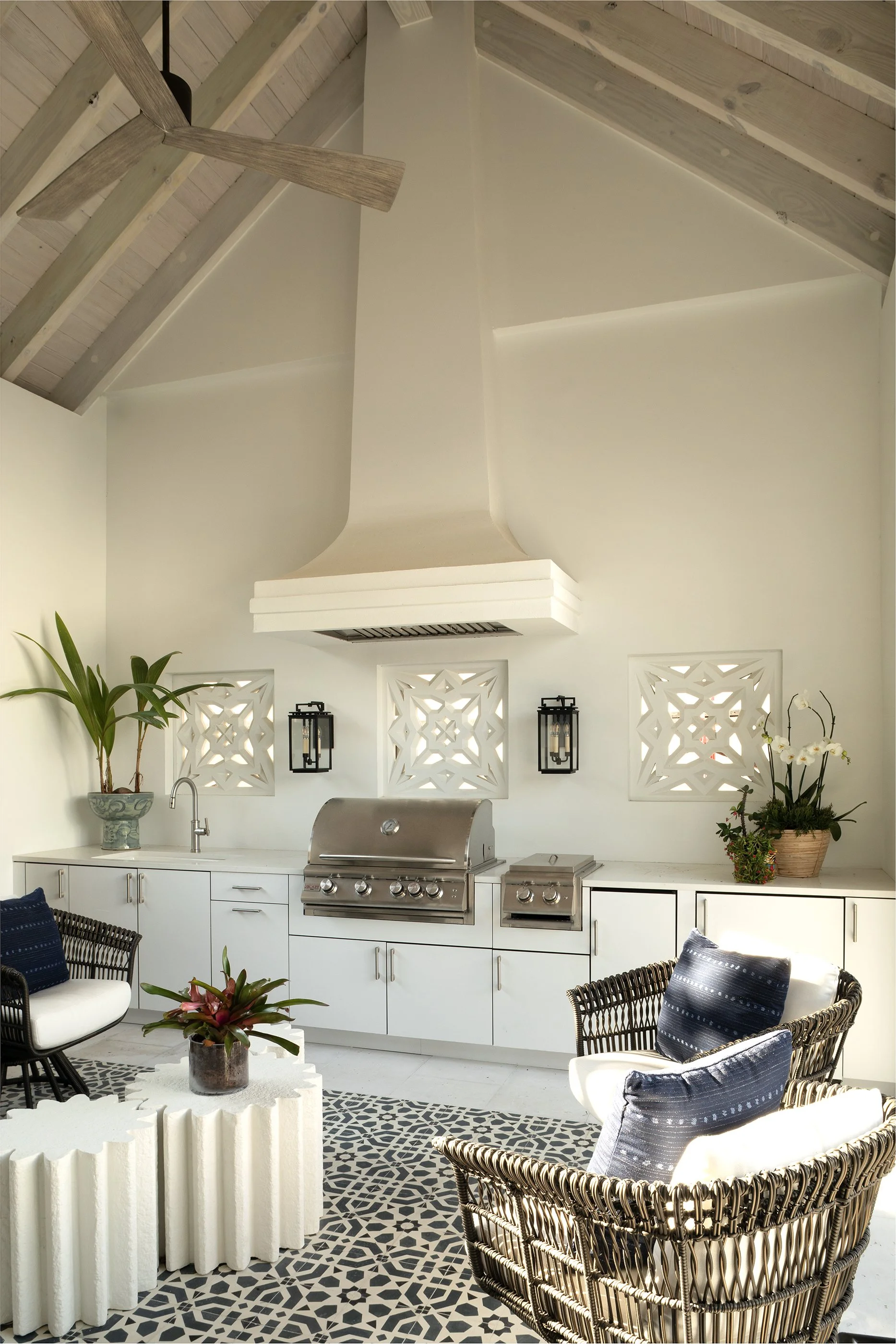 An outdoor kitchen with a ceiling fan, white cabinetry, stainless steel grill, black lantern lights, and black rattan seating with white and blue pillows, decorated with potted plants and a patterned area rug.