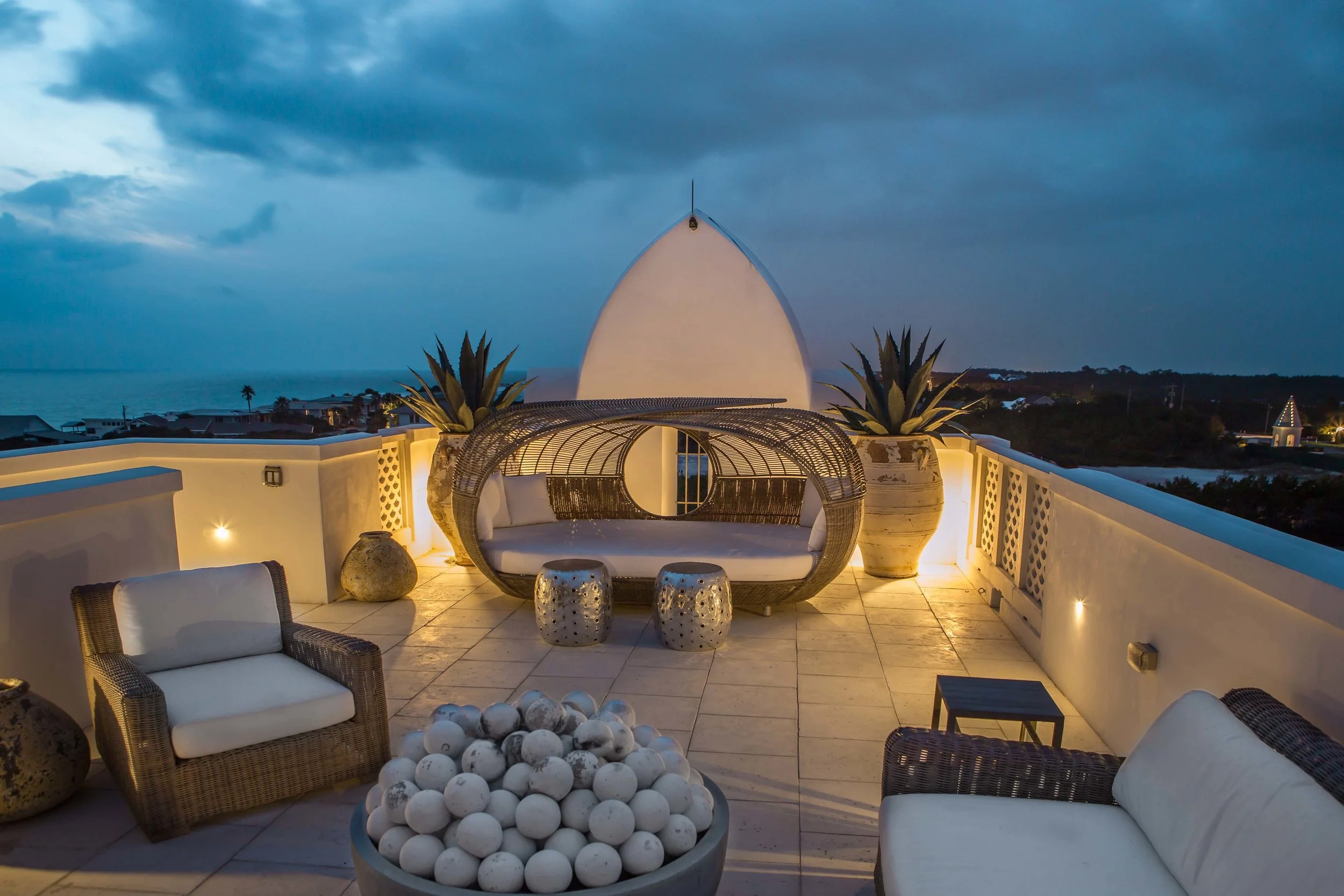 Rooftop terrace with outdoor seating and a view of the ocean at dusk, featuring wicker furniture, potted plants, and decorative white balls.