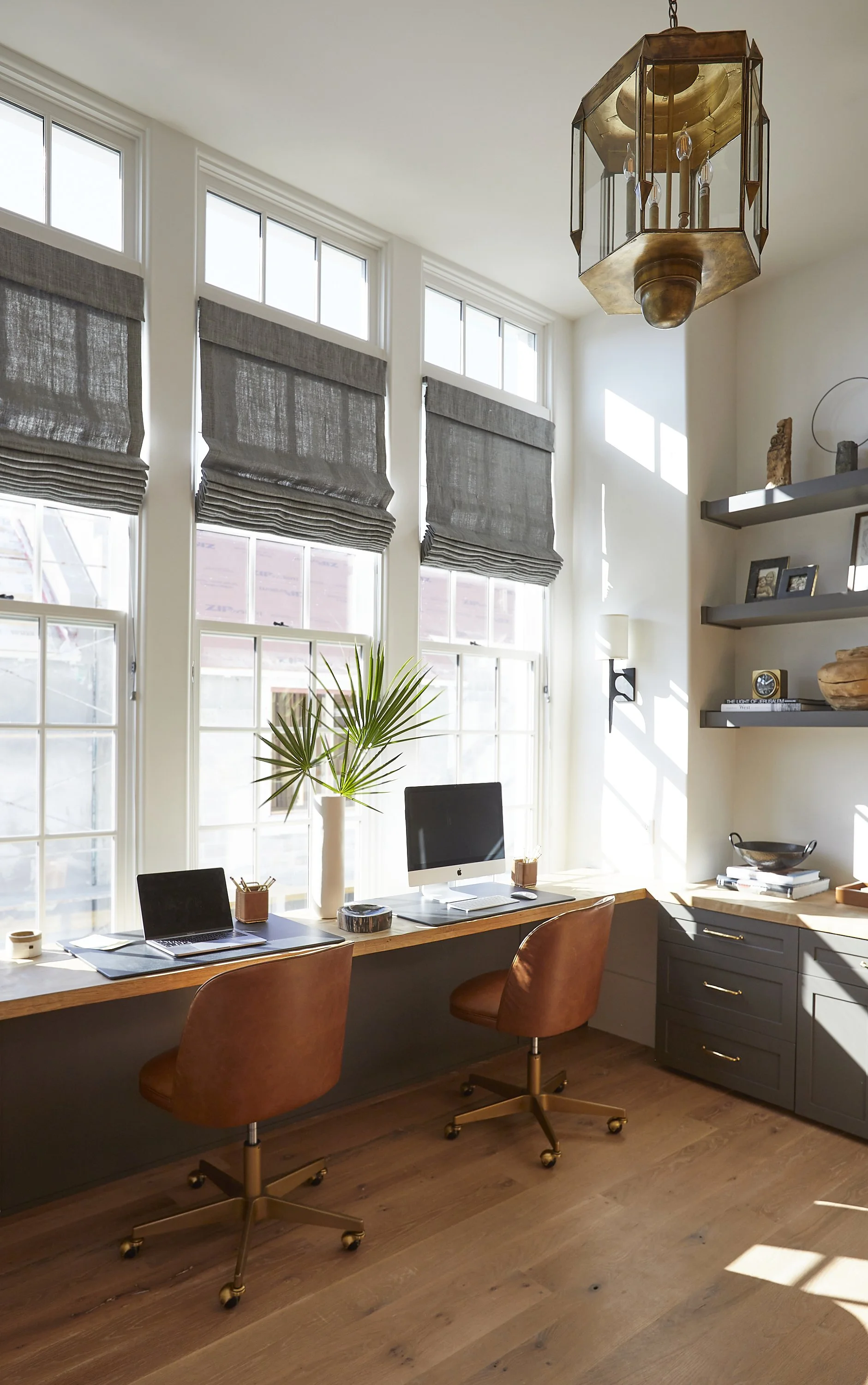 Bright home office with a long wooden desk against large windows, two brown swivel chairs, a computer, a tall vase with palm leaves, and wall shelves with decor and framed photos.