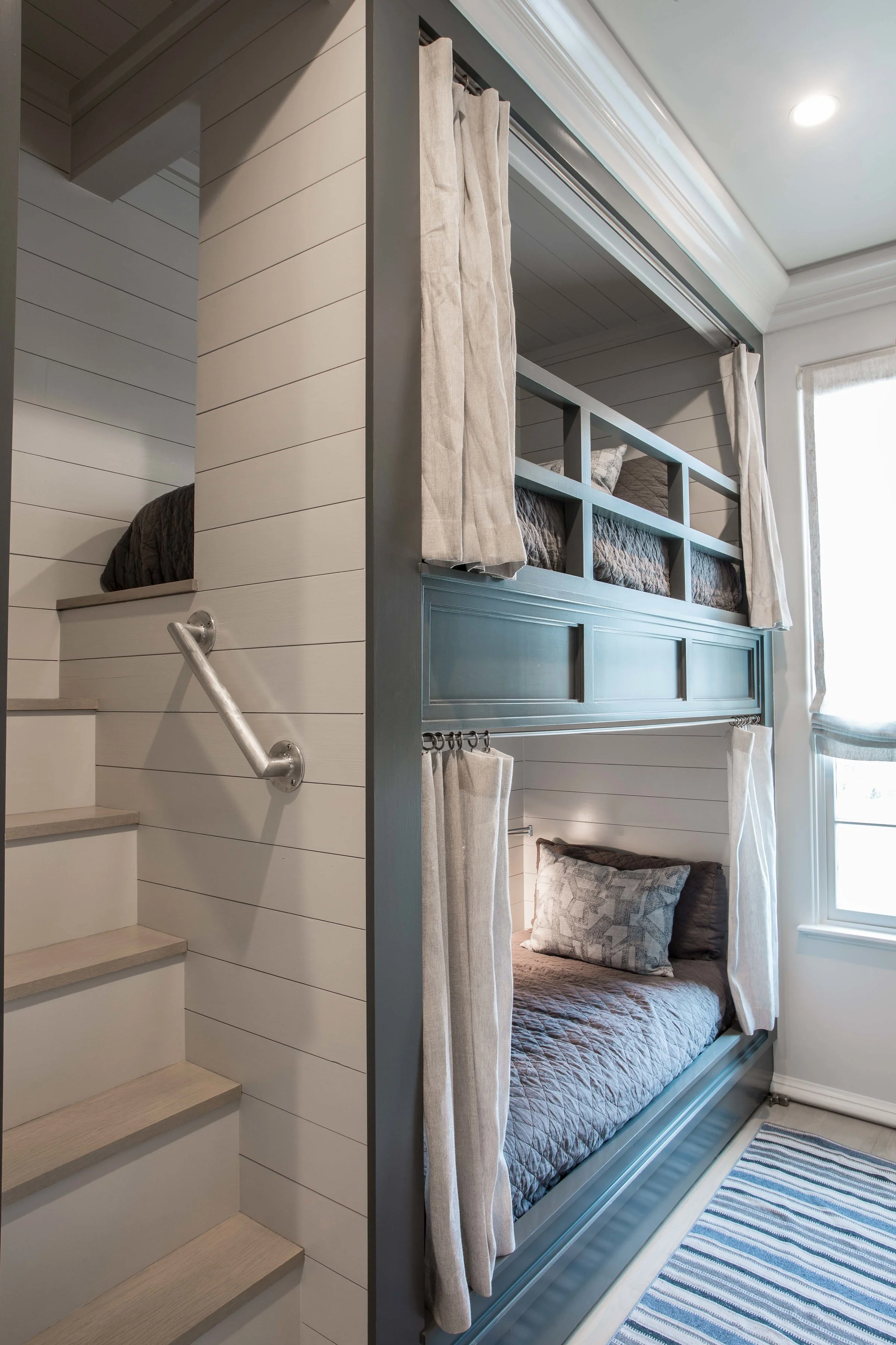 Bunk beds with curtains in a bedroom, adjacent to stairs with a handrail, white paneled walls, a striped rug, and a window with curtains.