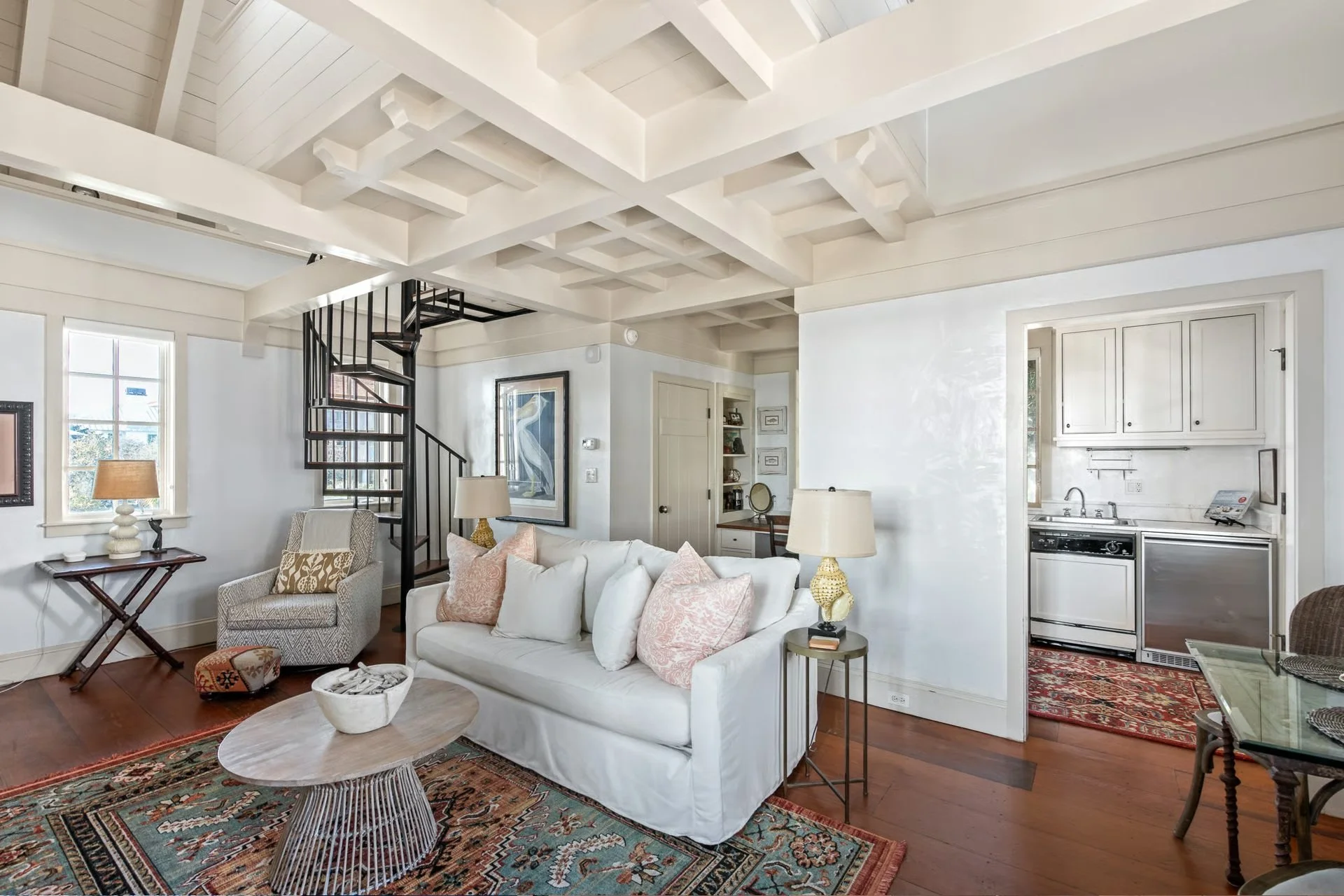 Living room with white sofa, armchair, and wooden coffee table, decorated with pillows and lamps. Spiral staircase leading to upper floor, and a small kitchen area with cabinets and appliances visible in the background.
